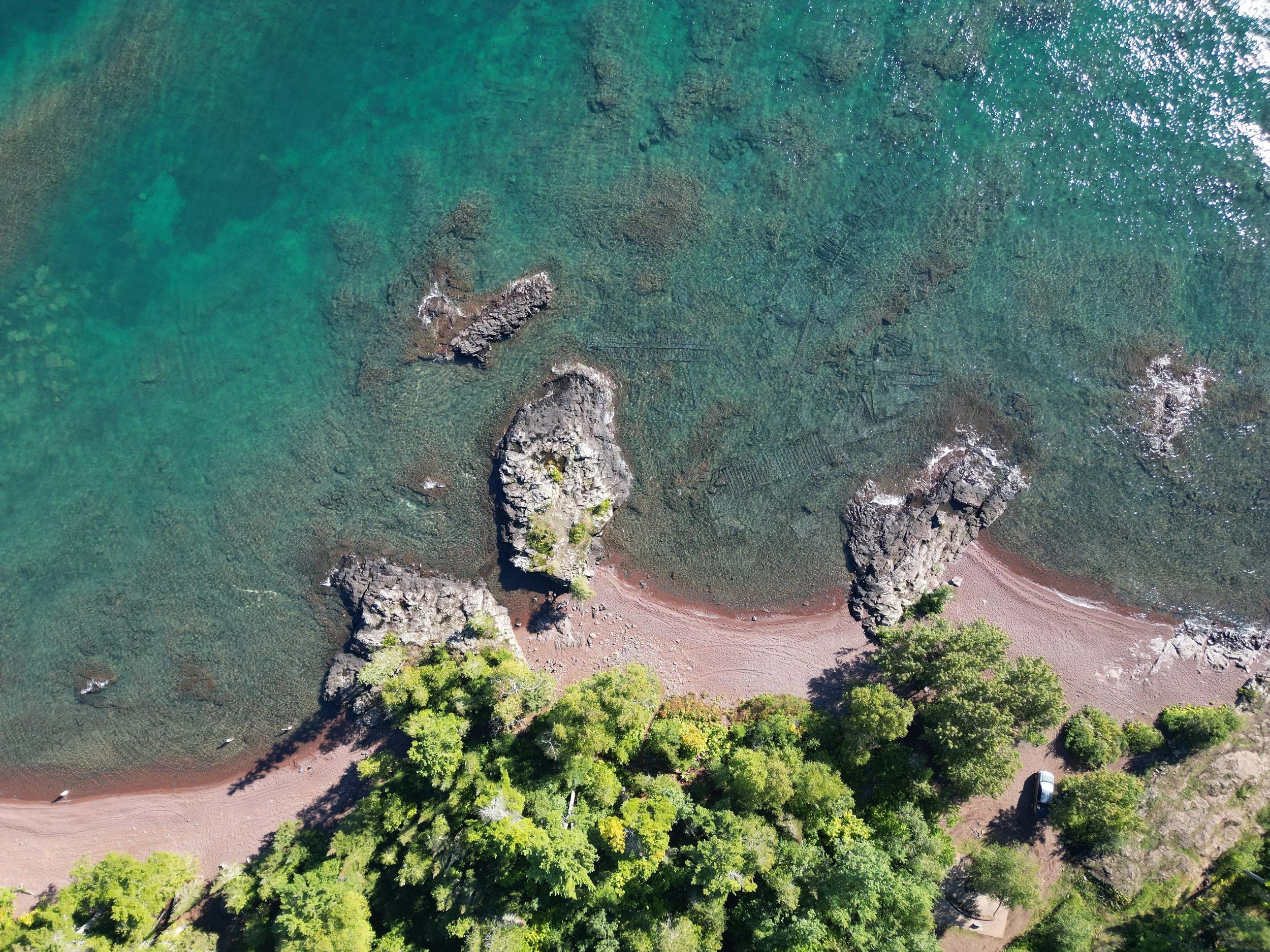 An aerial view of a coastal area with a sandy beach, green trees, and clear blue water with rocks and some waves.