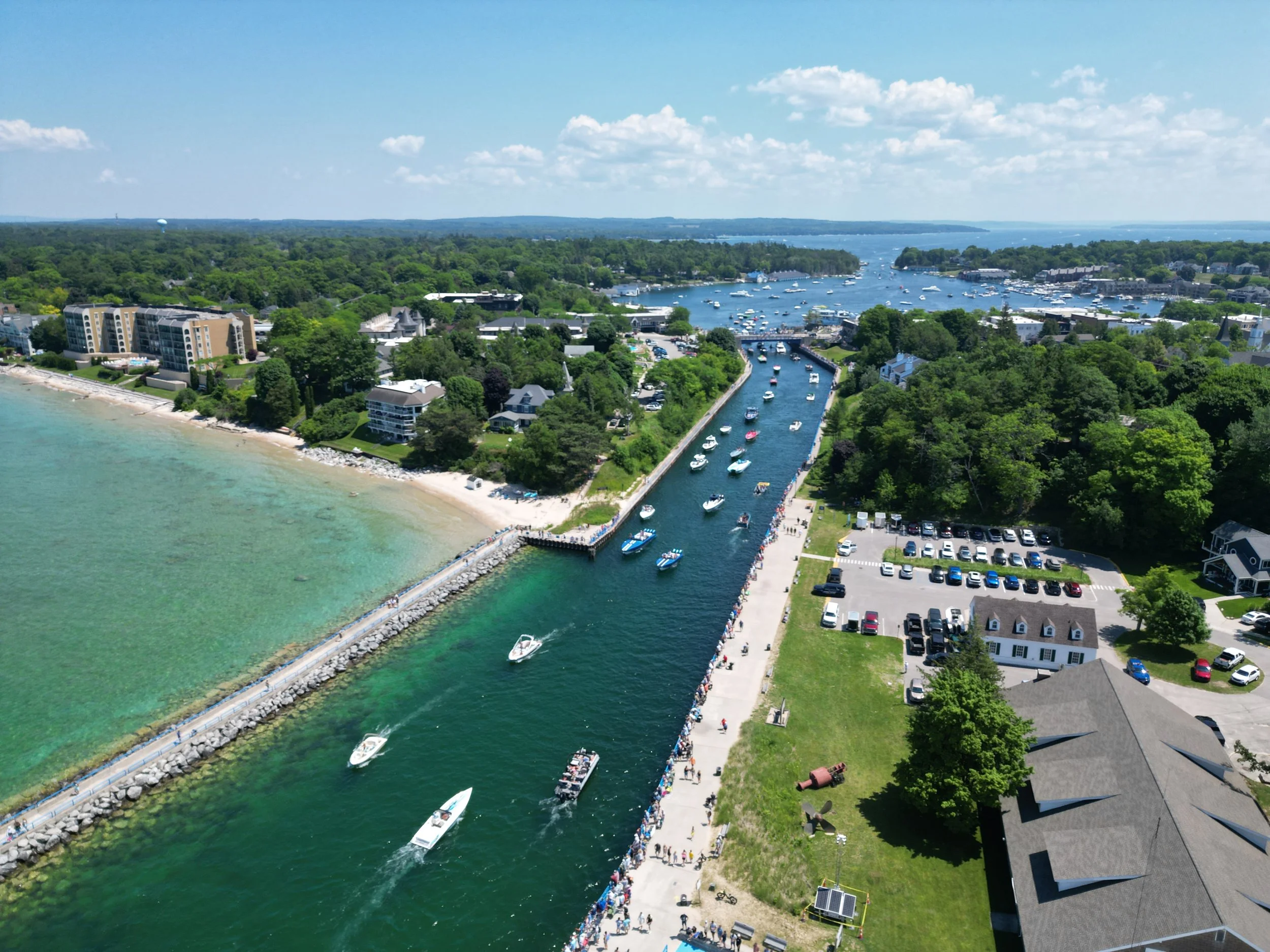 A coastal town with buildings, a marina filled with boats, a breakwater, and a crowded sidewalk with people enjoying a sunny day.