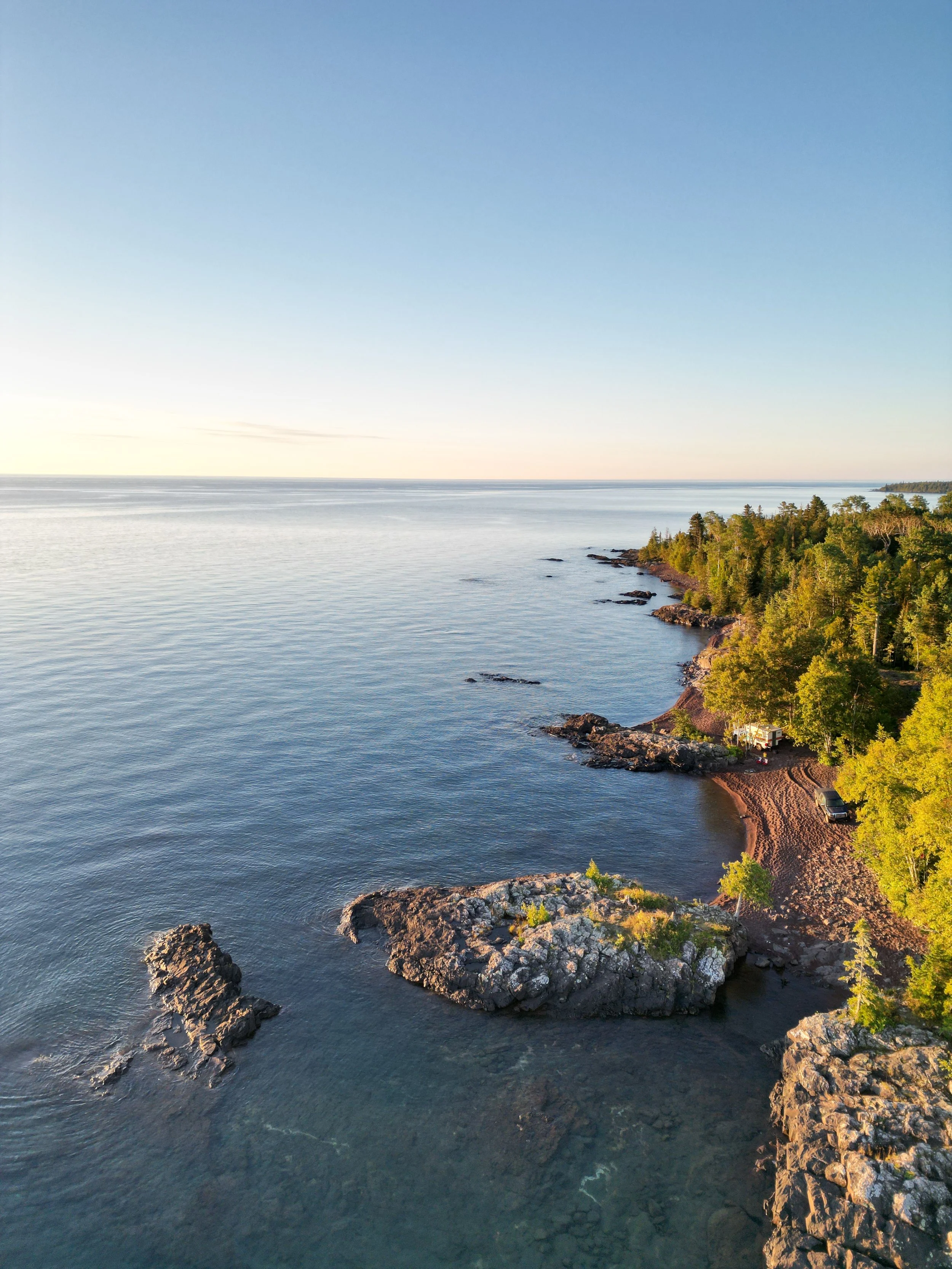 A serene coastal landscape with a rocky shoreline, calm water, and a forested area with a small vehicle parked near the trees.