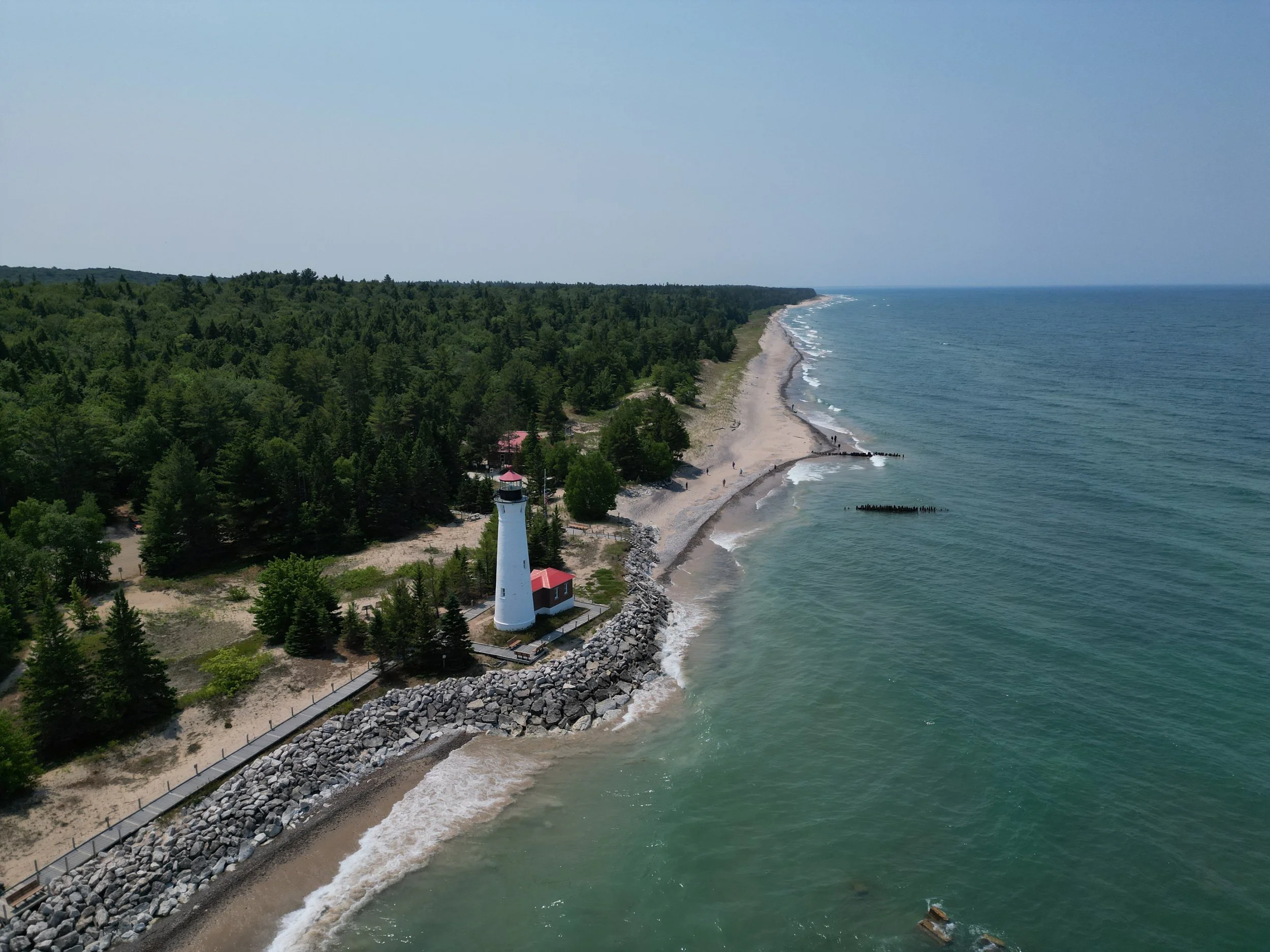 A lighthouse near the shore with a rocky barrier, surrounded by dense green trees, extending into the ocean on a clear day.