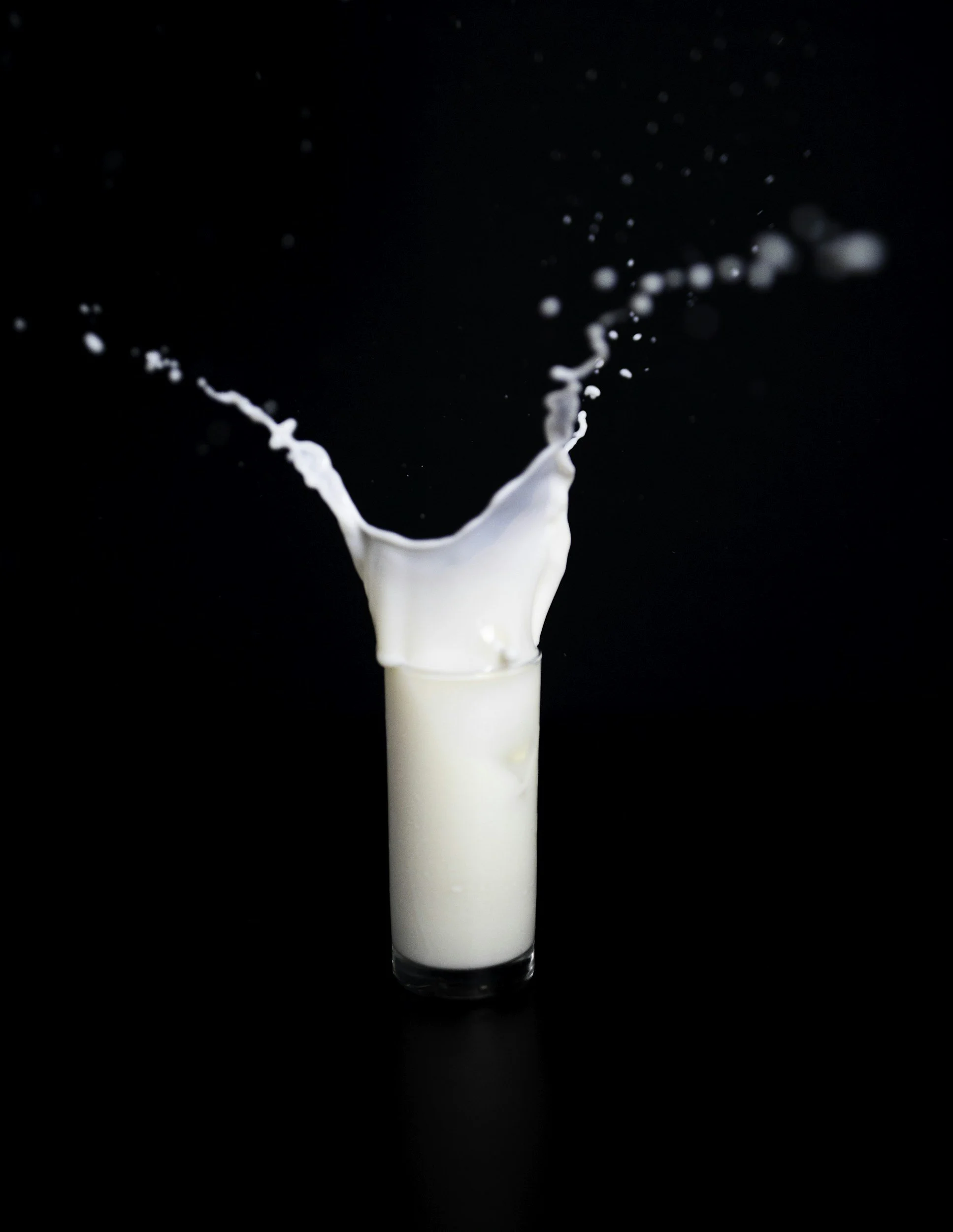 A glass of milk being poured with milk splashing out against a black background.