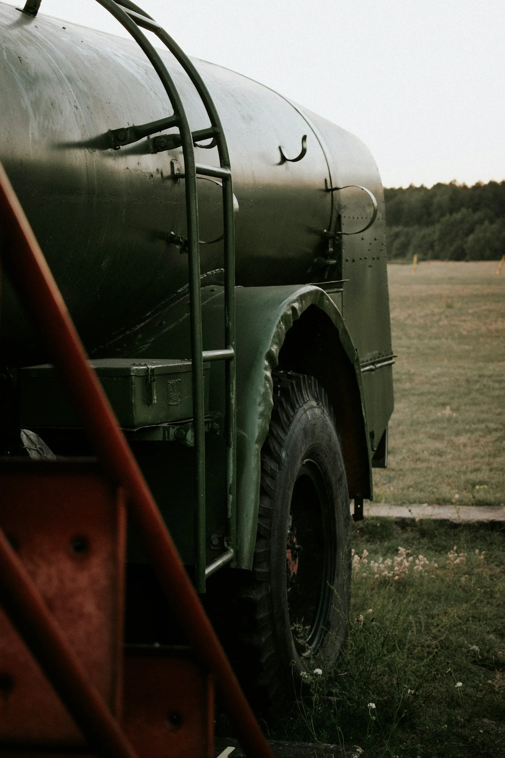 Close-up of a military vehicle with a black, rounded body, large tires, and a rear hatch, parked on a grassy field during dusk with a background of trees and an open sky.