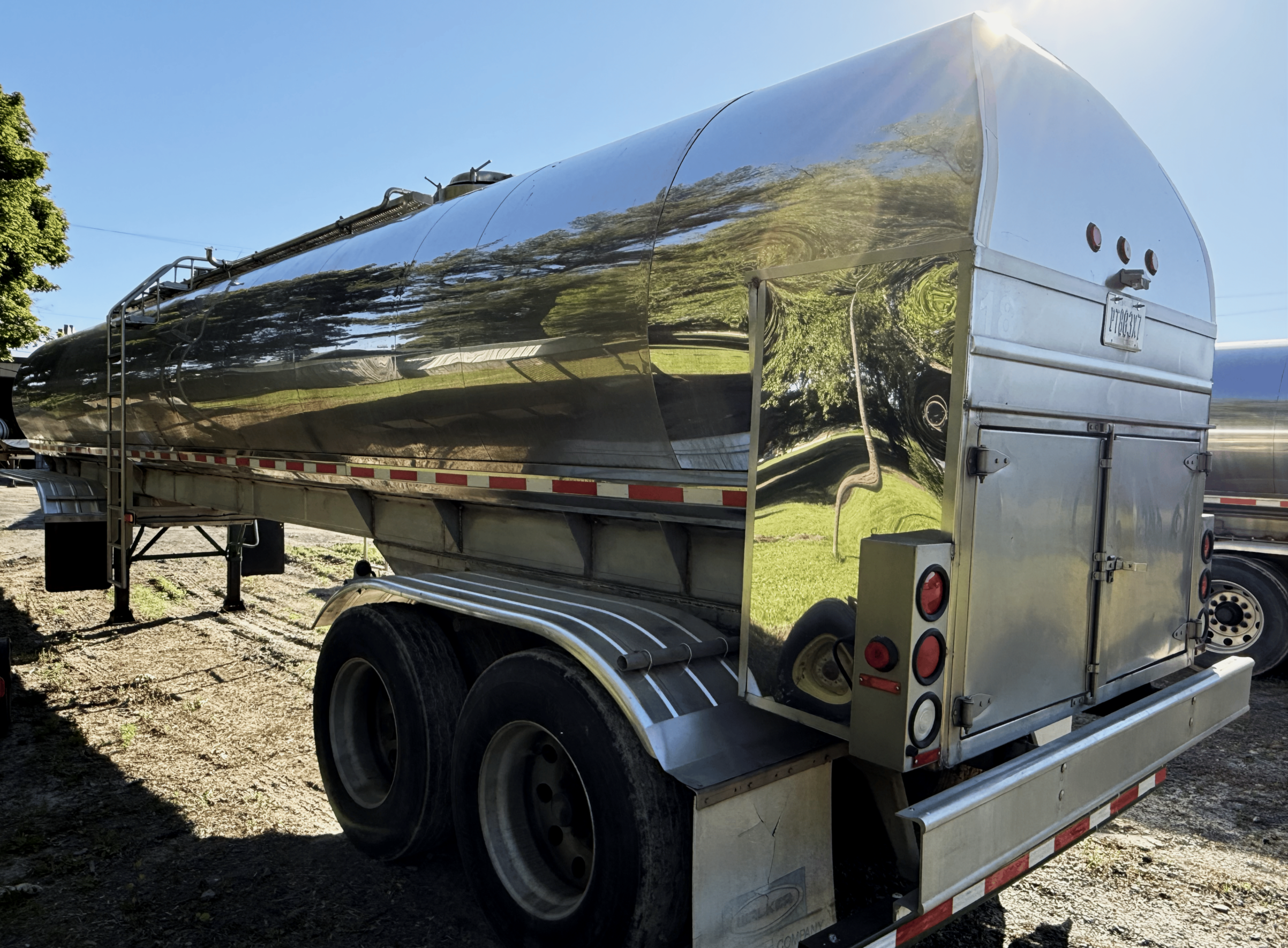 A shiny, silver, cylindrical trailer with a rounded front parked outdoors on a sunny day. The trailer's surface reflects the surrounding trees and grass. It has black tires, metal steps, and red and white safety reflective tape along the bottom.