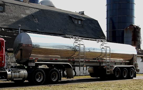 A large shiny metal food grade farm pickup tanker trailer air ride suspension parked on grass next to buildings.