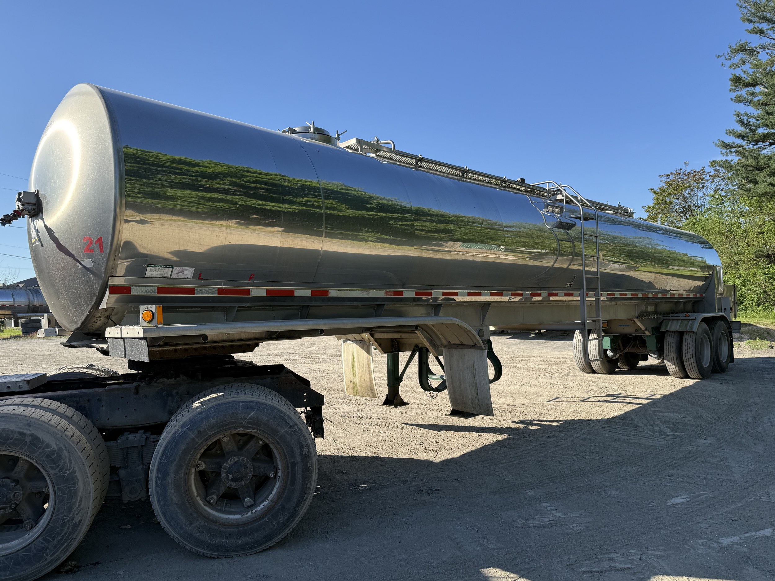 A shiny silver food grade farm pickup tanker trailer air ride suspension parked on a dirt surface with green trees and a clear blue sky in the background.