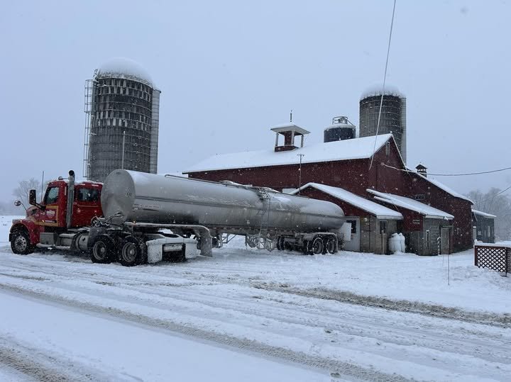 A red barn and a tanker truck in a snowy farm setting with two grain silos in the background.