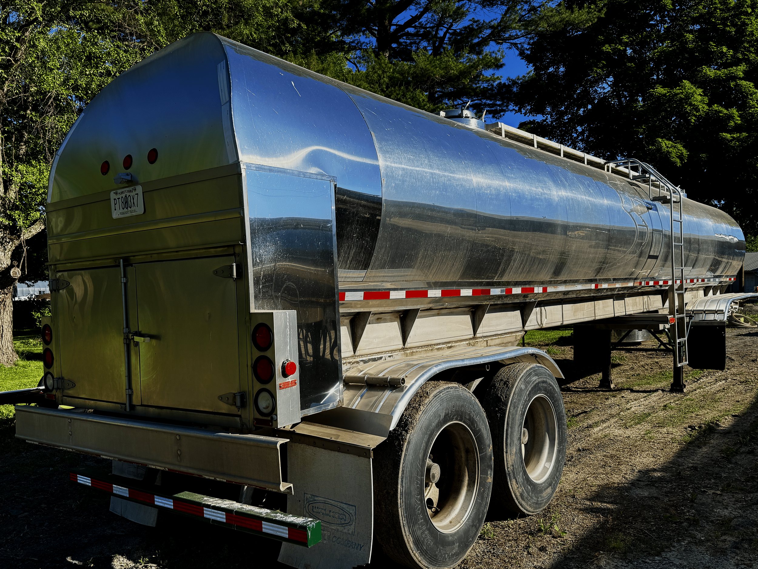 A shiny, metallic silver food grade farm pickup tanker trailer air ride suspension attached to a truck, parked outdoors with trees and a clear blue sky in the background.