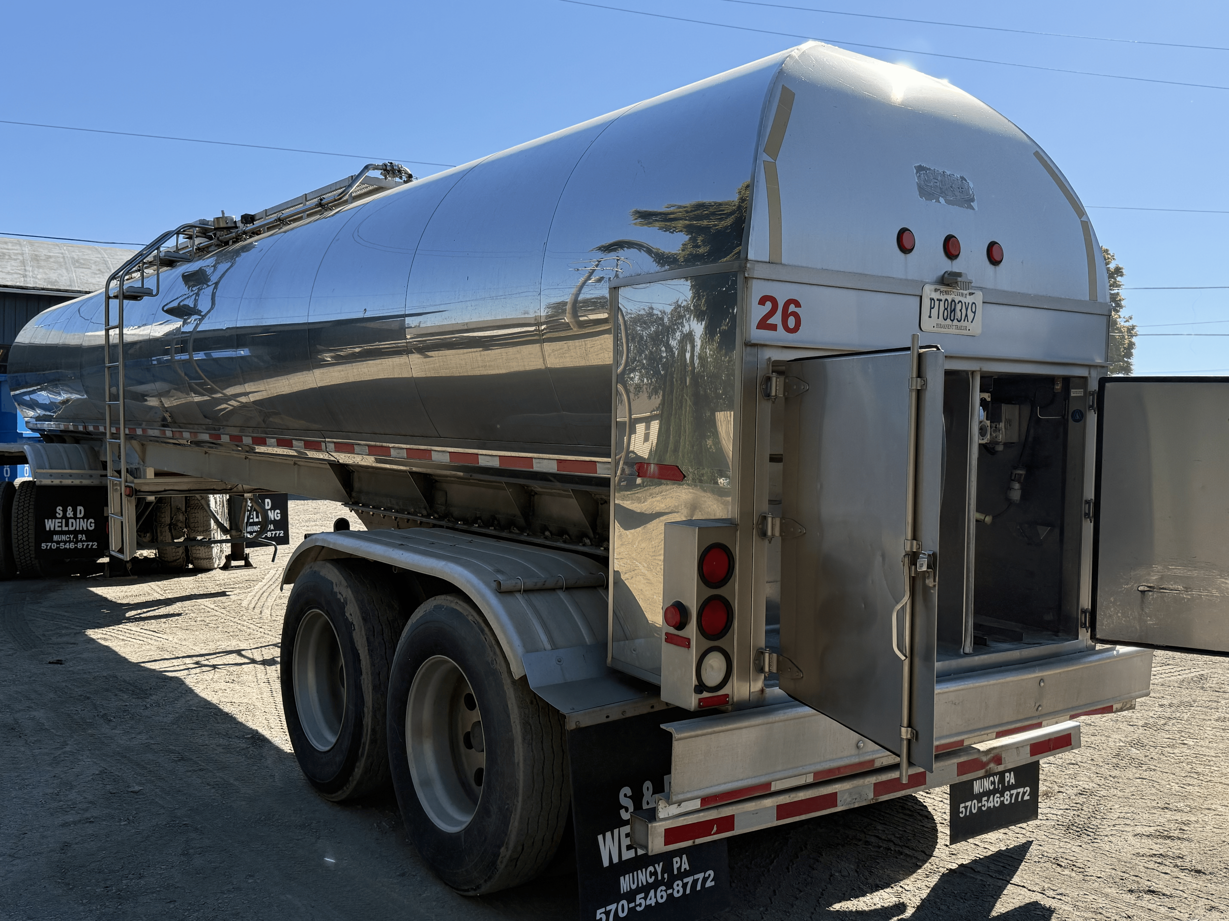 A shiny metal tanker truck parked outdoors on a clear day, with a blue sky, trees, and power lines in the background. The truck has a Pennsylvania license plate and signage for a welding company in Muncy, PA.
