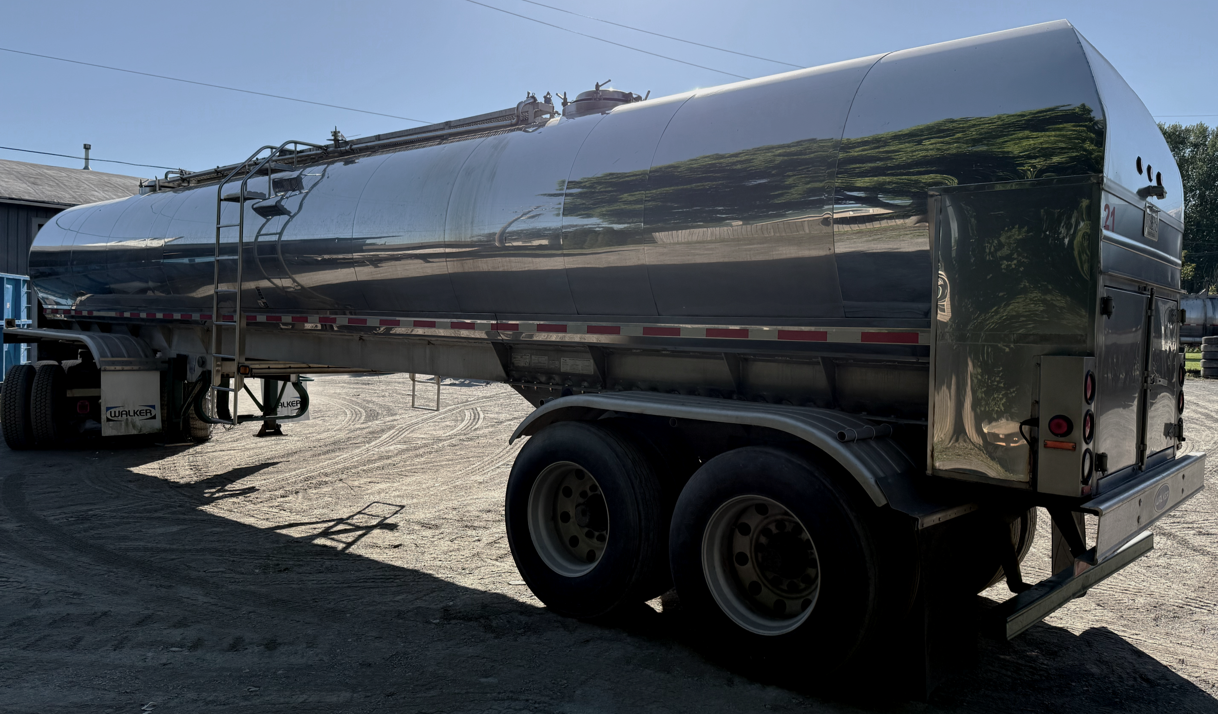A shiny stainless steel food grade farm pickup tanker trailer air ride suspension parked on a gravel lot with a building and trees in the background.