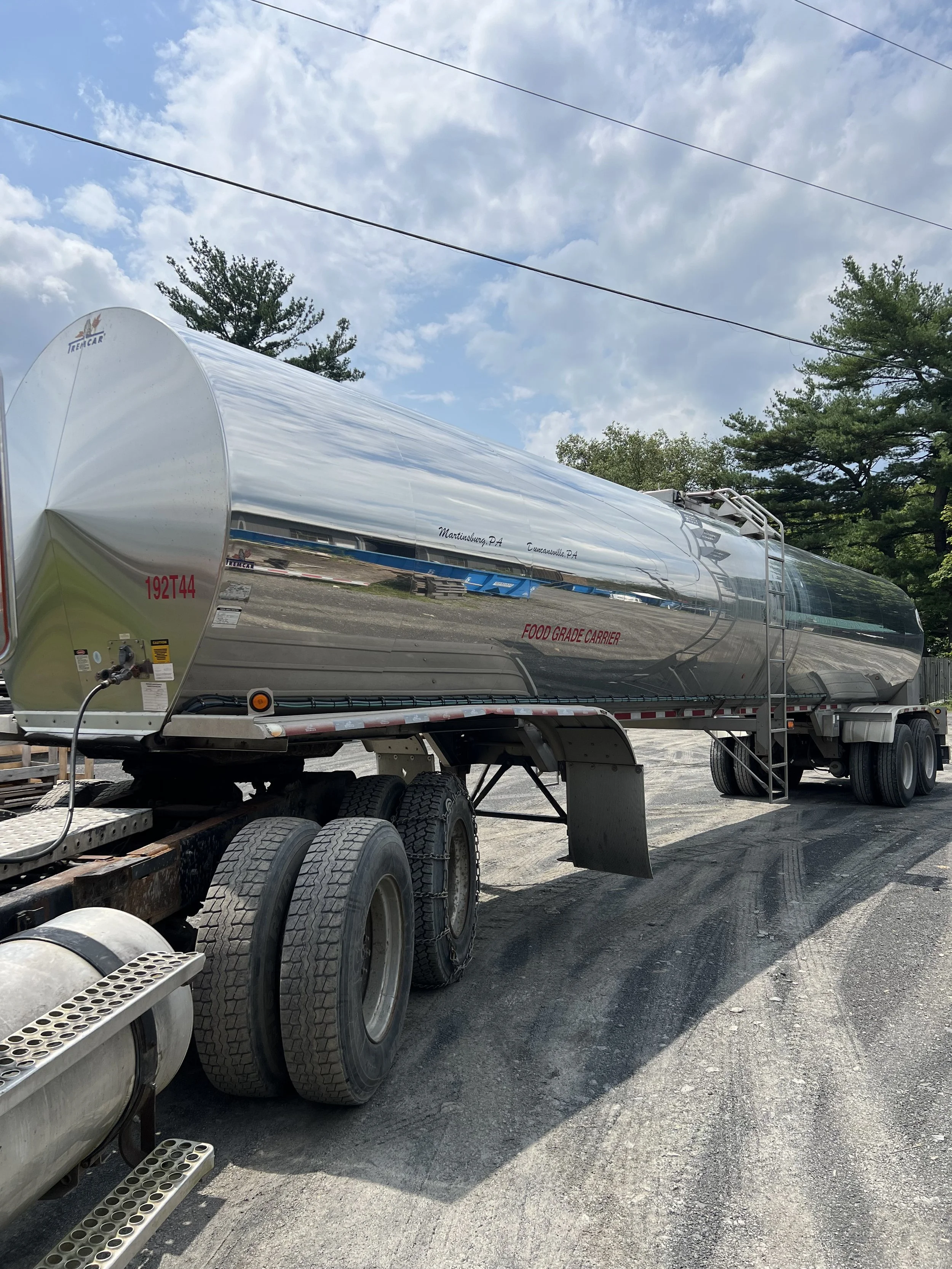 A large, shiny, silver  food grade farm pickup tanker trailer air ride suspension parked outdoors on a gravel surface, with a partly cloudy sky and trees in the background.