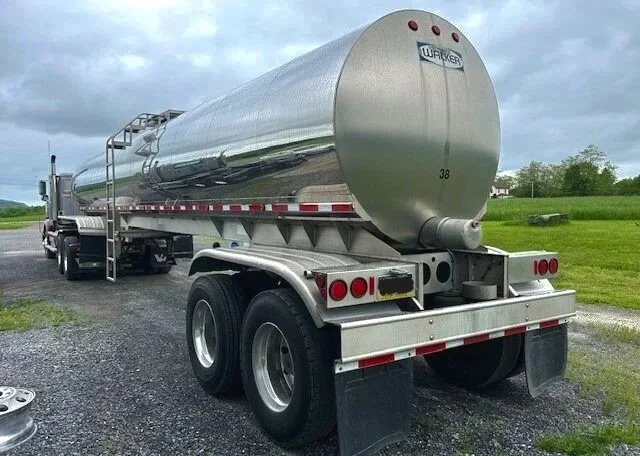 A large stainless steel food grade farm pickup tanker trailer air ride suspension parked outdoors on a gravel surface with green grass and cloudy sky in the background.