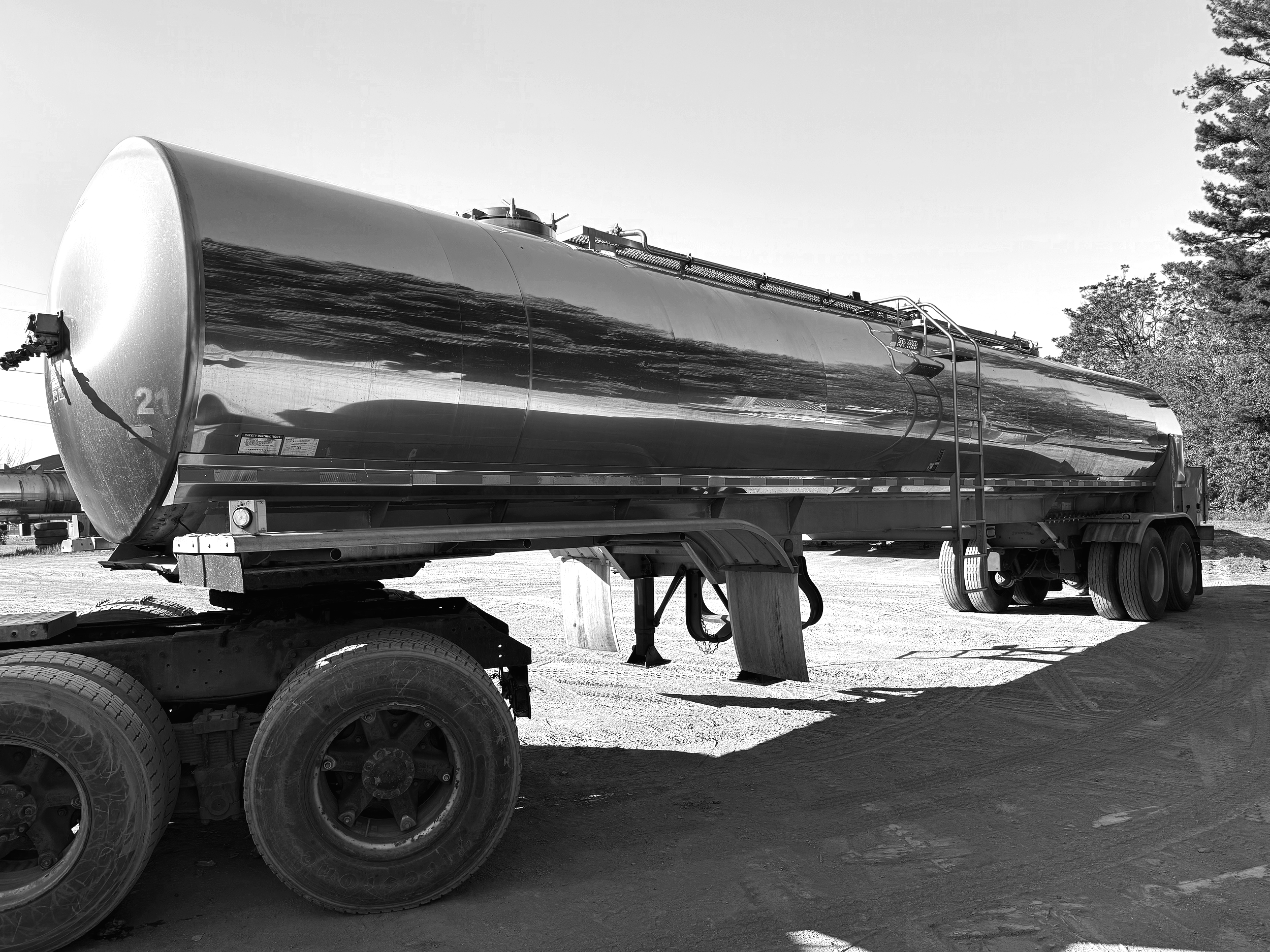 A large cylindrical metal tanker truck parked outdoors with trees in the background, black and white photograph.