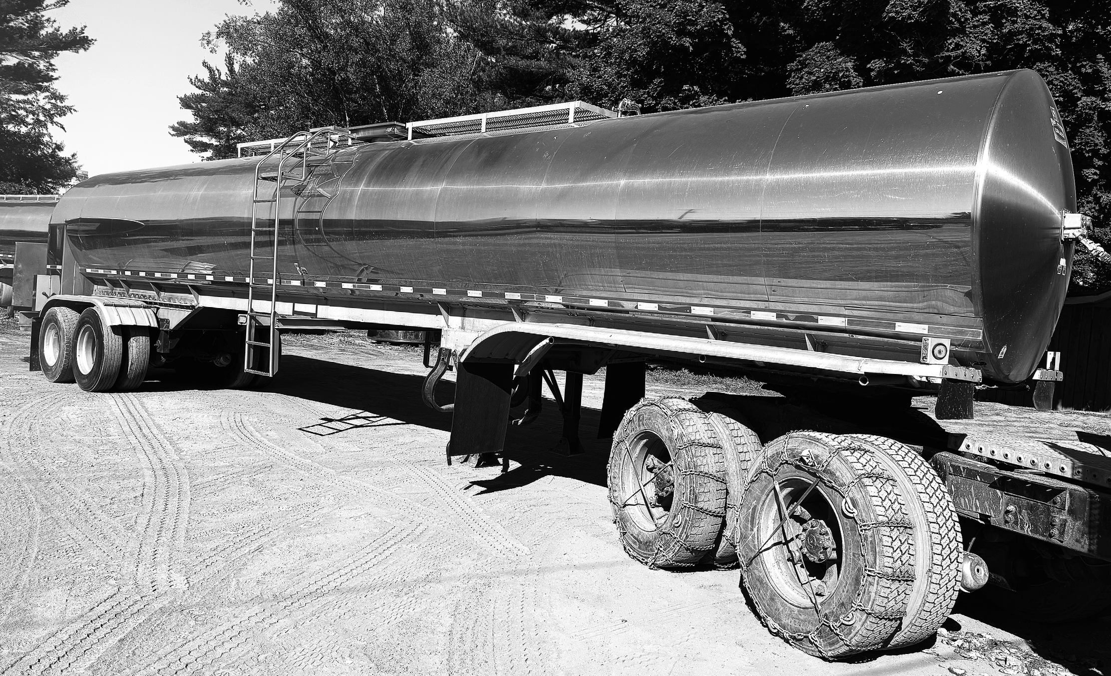 A large cylindrical metal  food grade farm pickup tanker trailer air ride suspension mounted on a trailer truck with tires, parked on a dirt ground, with trees in the background.