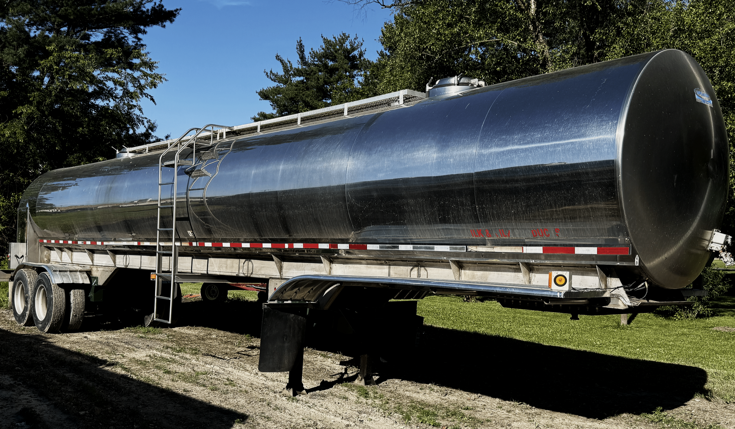 A large, shiny metal food grade farm pickup tanker trailer with air ride suspension parked on grass with trees and blue sky in the background.