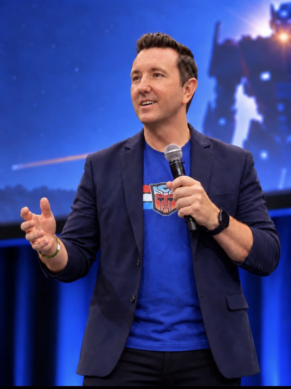 Dr. Peter Reiley speaking about leadership and pop culture at an event, holding a microphone, wearing a blazer over a blue T-shirt with an Autobot logo from Transformers, standing in front of a space-themed backdrop with Optimus Prime.