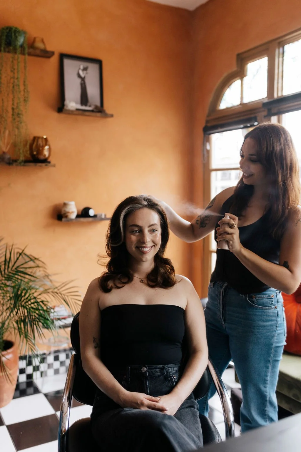 A woman getting her hair styled with hairspray by another woman in a cozy, well-decorated room.