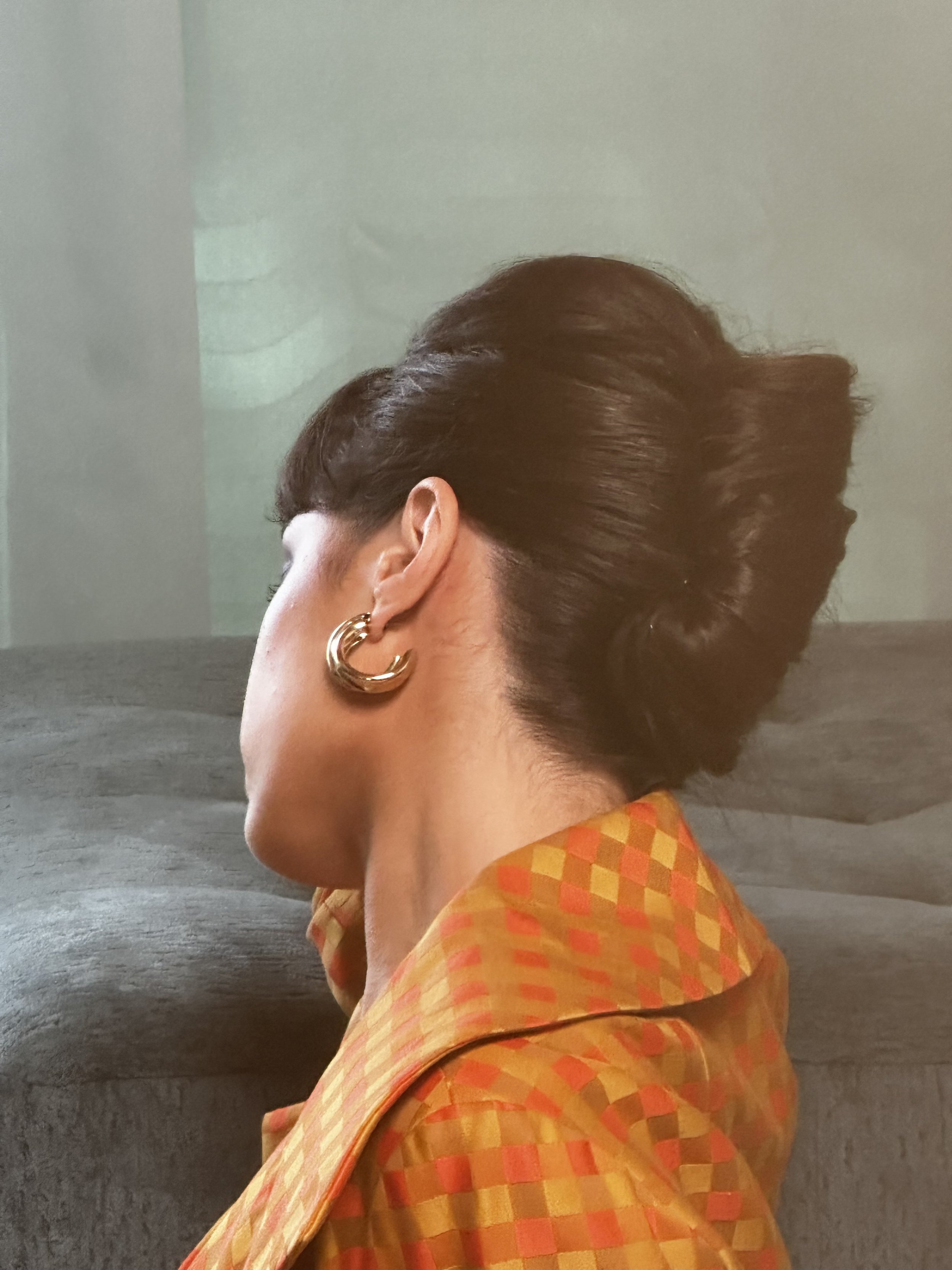 Close-up side profile of a woman with short, wavy brown hair, wearing a gold hoop earring and an orange checkered blouse, against a dark background.