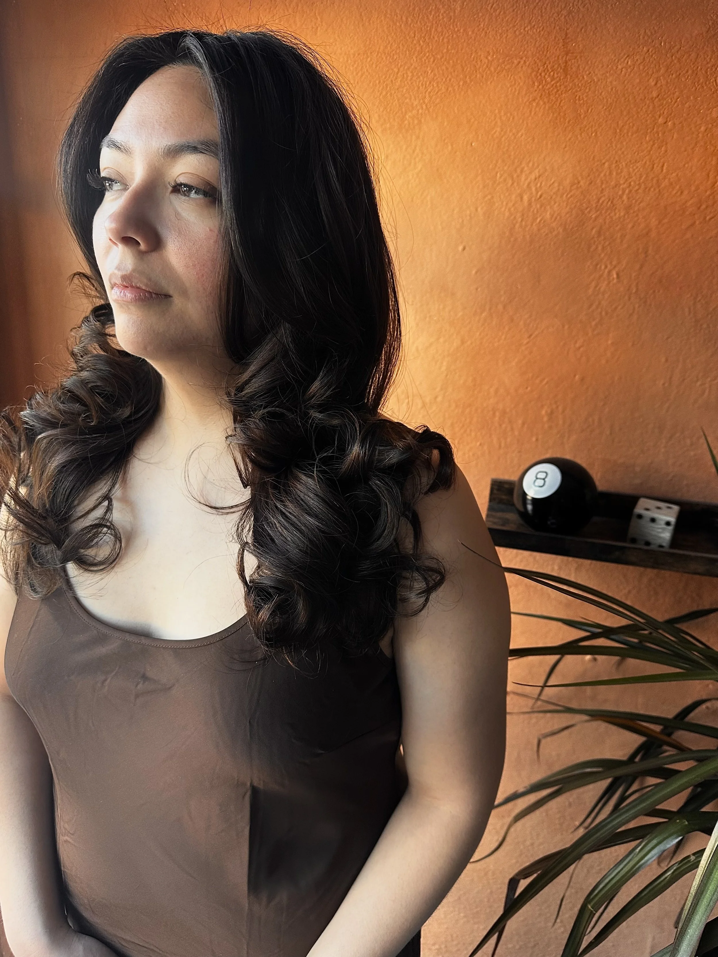 A woman with dark curly hair and light skin, wearing a brown sleeveless top, is facing left. She appears to be indoors against an orange wall, with a black and white object and some plant leaves visible in the background.