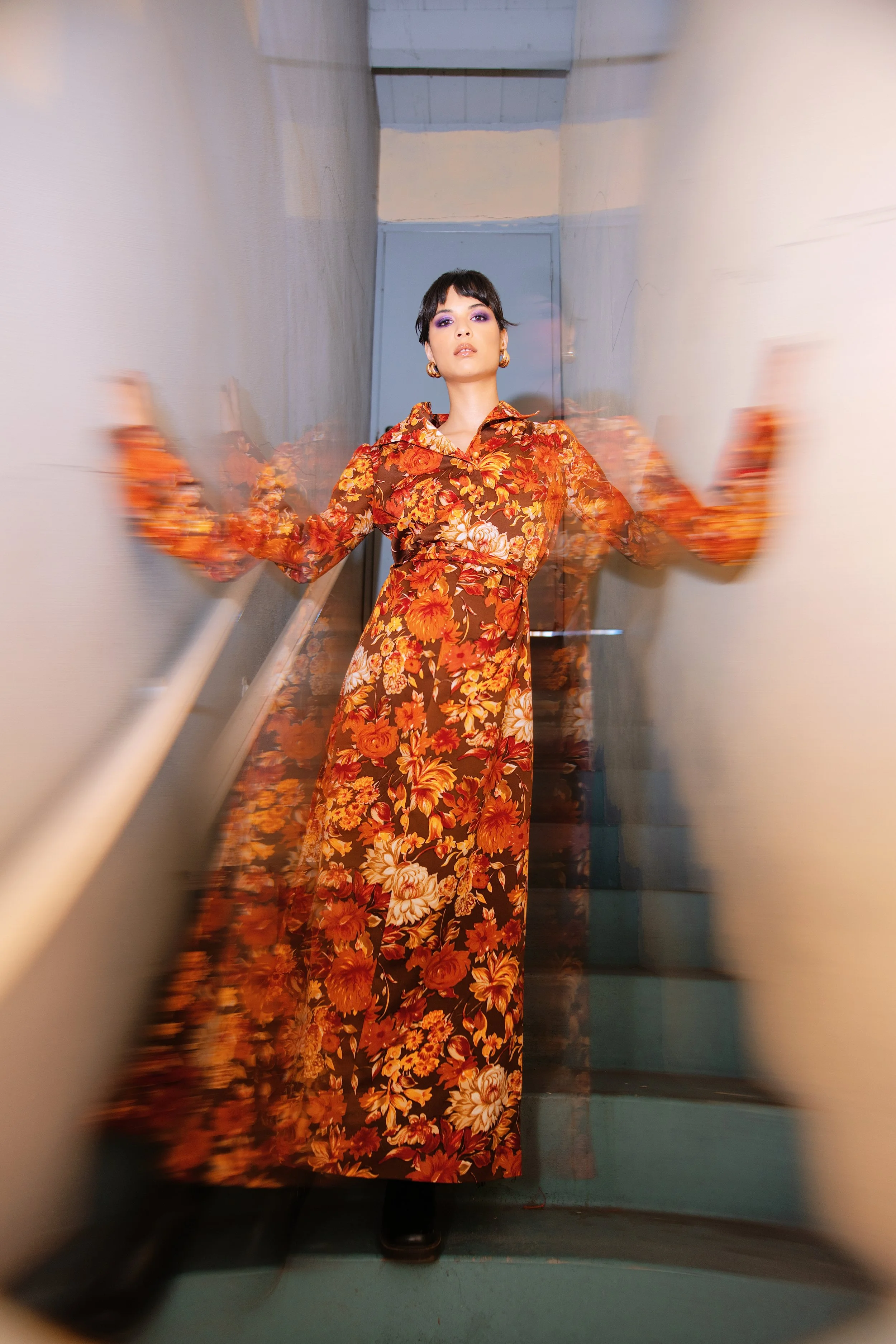 A woman with short dark hair, wearing a long floral dress with orange, red, and beige flowers, is standing in a narrow, slightly blurry hallway with light-colored walls and blue wooden ceiling beams.