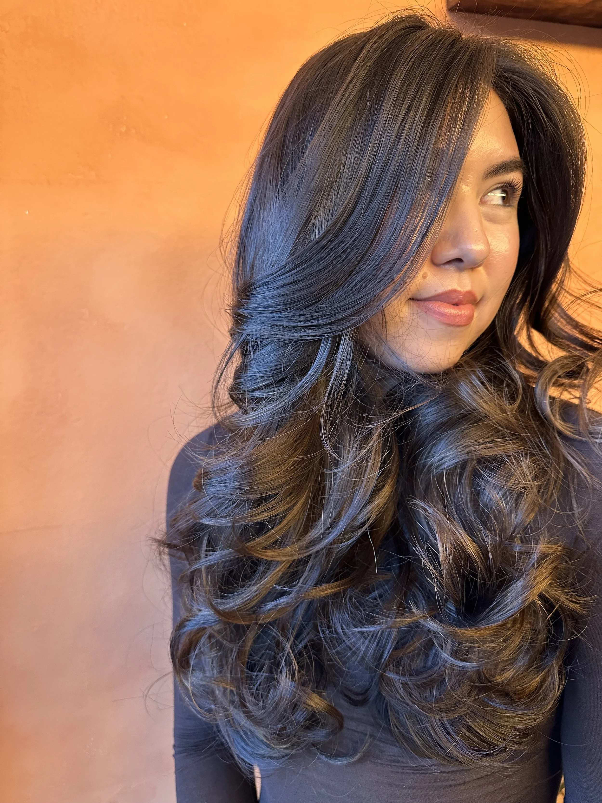 A woman with long hair and a bouncy blowout, sitting against a reddish-orange wall, looking to the side with a slight smile.