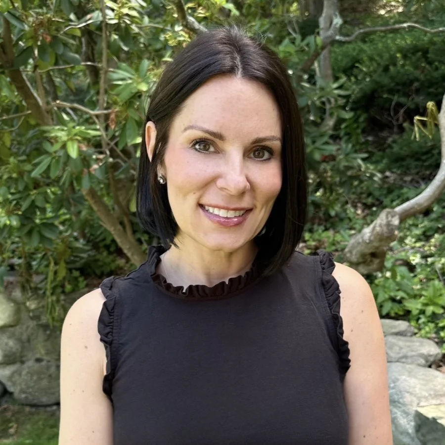 A woman with shoulder-length dark hair, smiling, wearing a black sleeveless top with ruffled collar and ruffled sleeves, standing outdoors with green bushes and trees in the background.