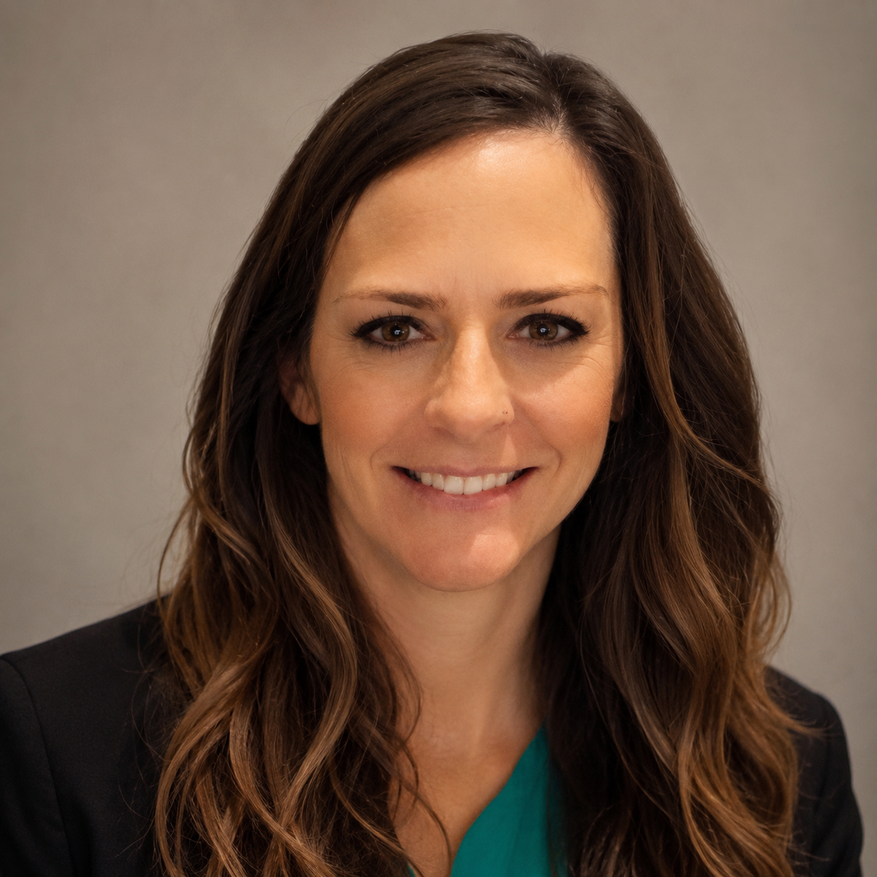 A woman with long, wavy brown hair and light skin, smiling at the camera, wearing a dark blazer over a teal top, against a neutral background.