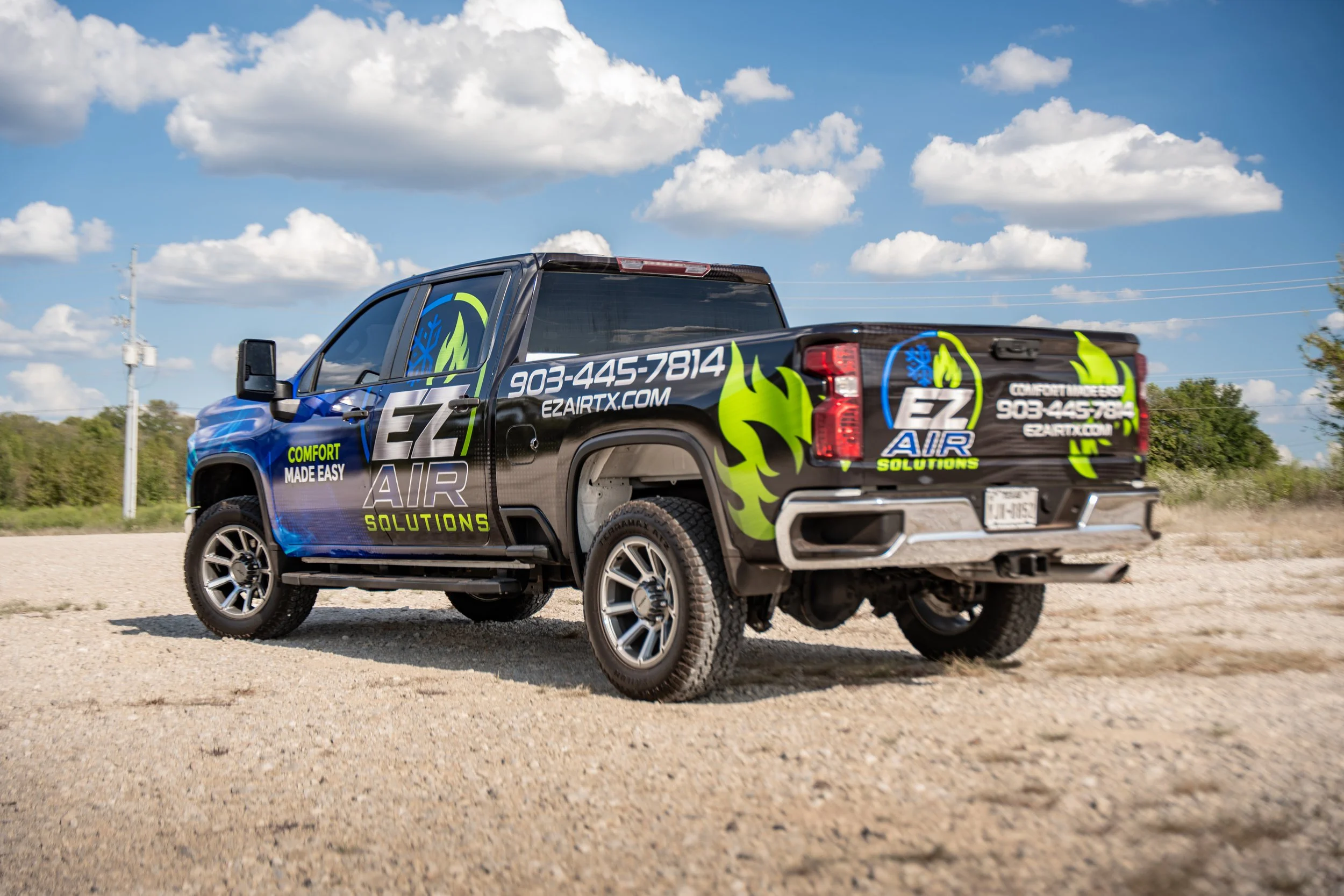 A black pickup truck with colorful branding for EZ Air Solutions drives on a gravel road under a partly cloudy sky.