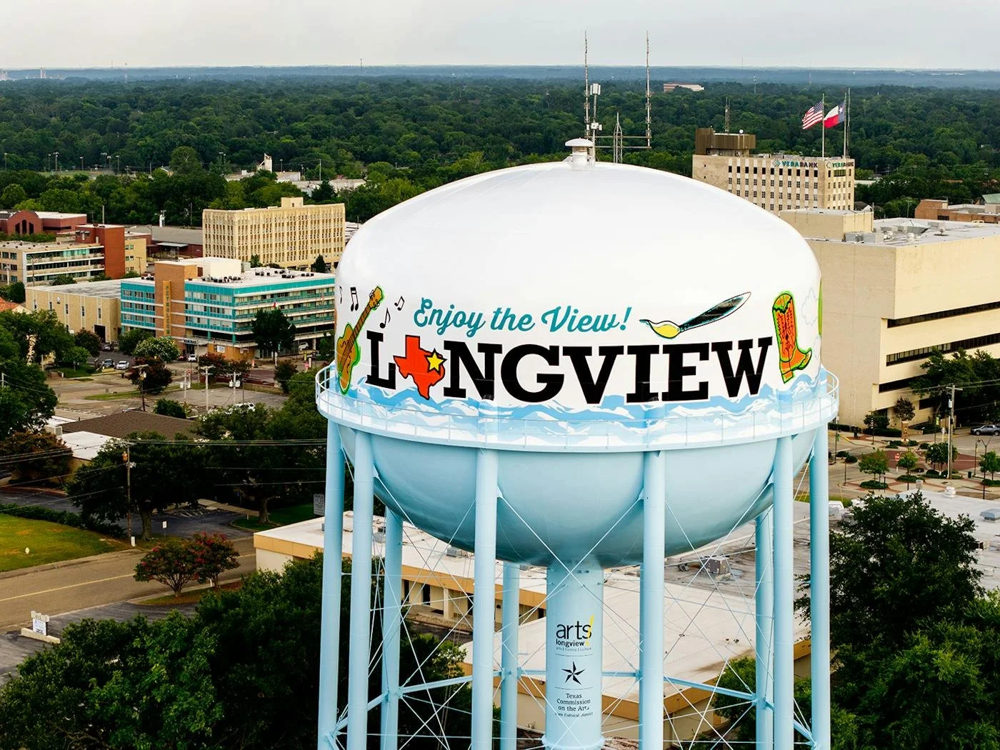 A large white water tower in Longview, Texas, with colorful graphics and the words 'Enjoy the View! Longview' painted on it. The tower is supported by multiple light blue structural supports, and the cityscape with trees and buildings is visible in the background.