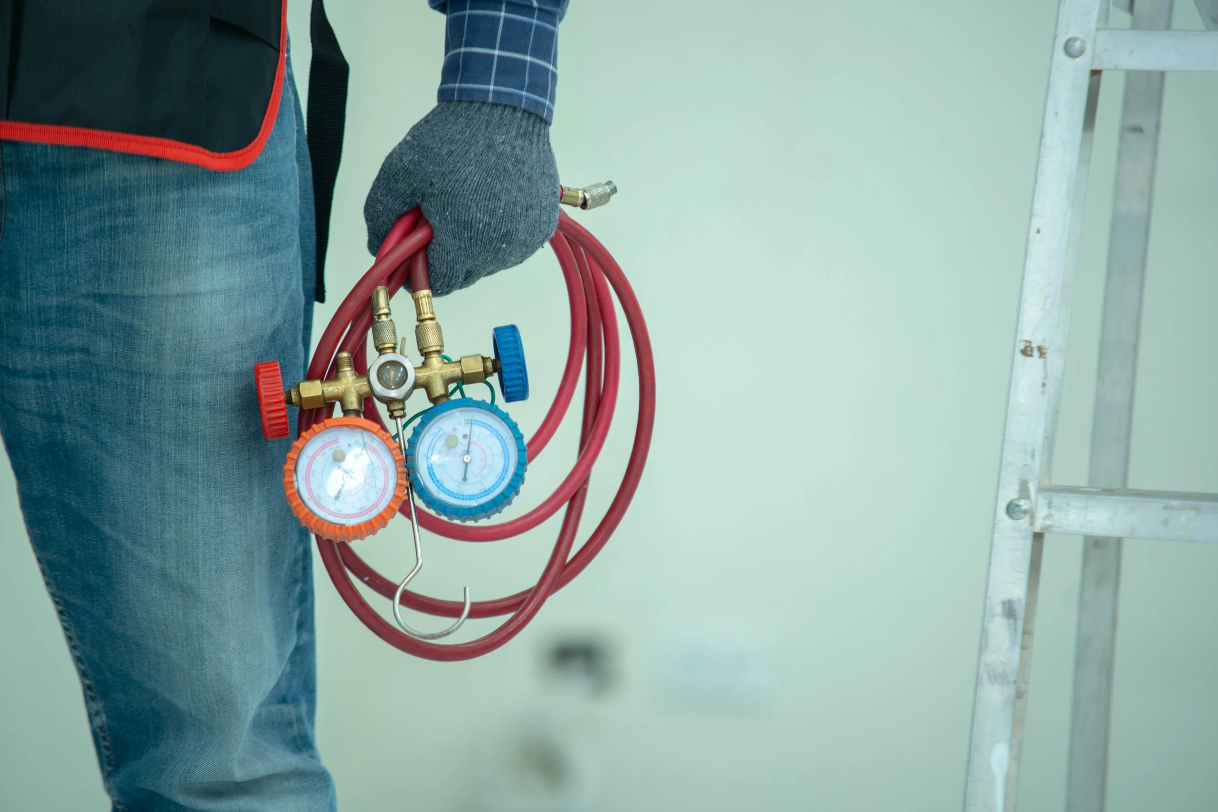 A person wearing a gray work glove holding red hoses and pressure gauges, with a ladder to the right side.