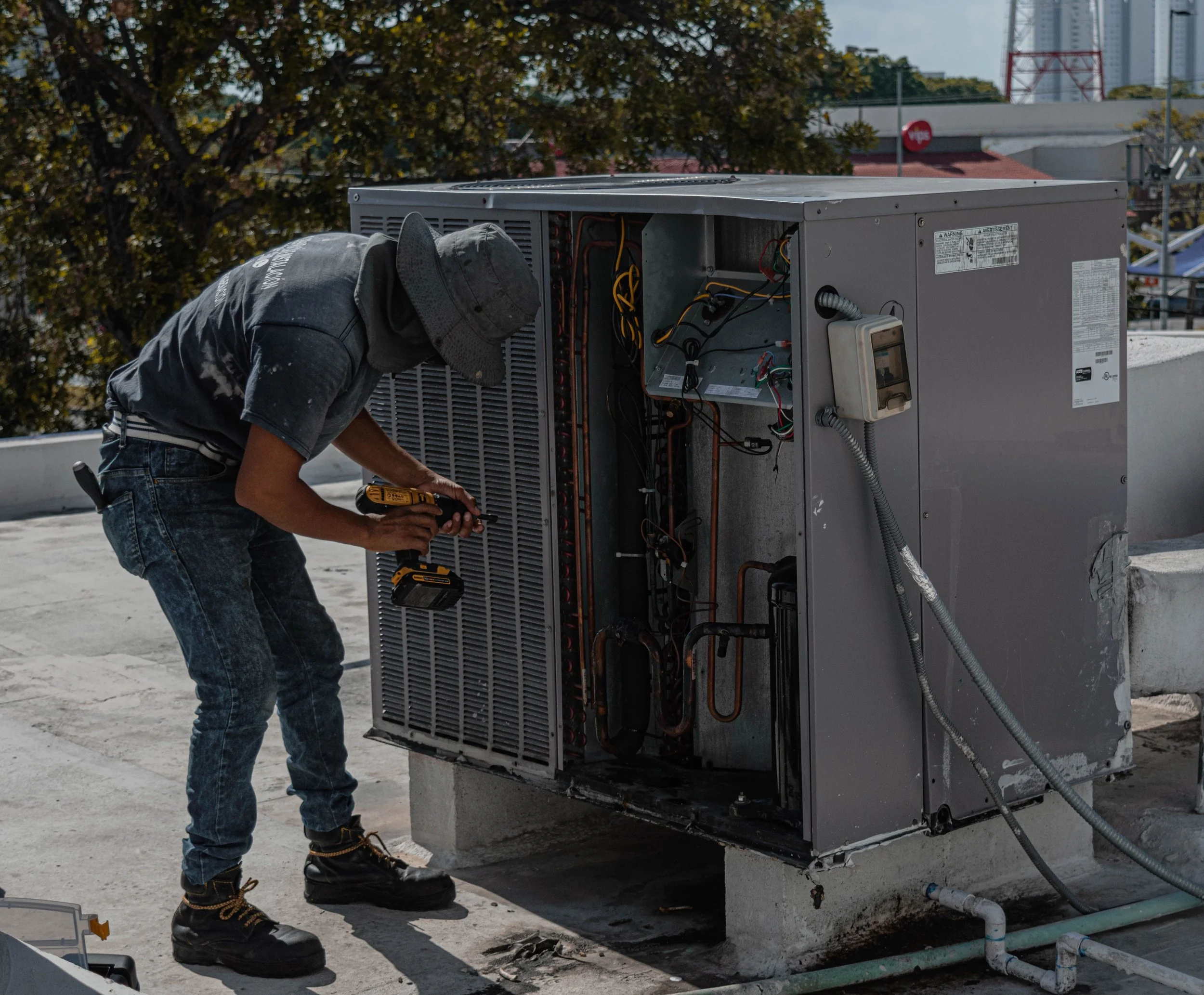 A technician working on a HVAC unit on a rooftop, using tools, with trees and city buildings in the background.