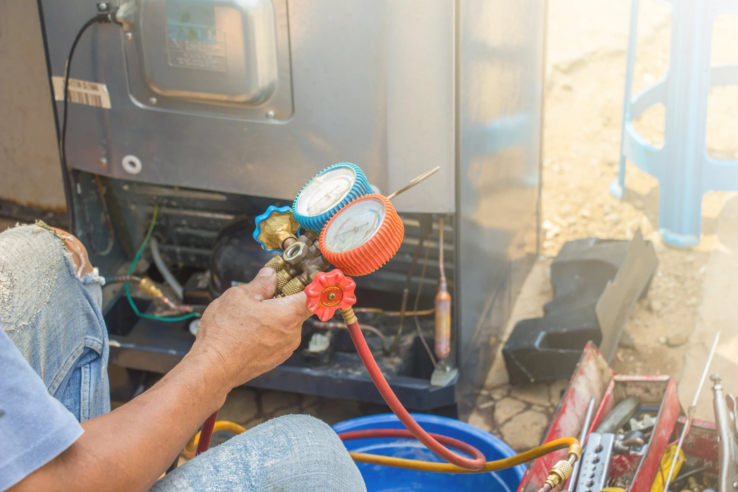 A person working with refrigeration gauges attached to an air conditioning unit or compressor, using a red hose and a pressure gauge, in an outdoor setting with construction tools and equipment nearby.