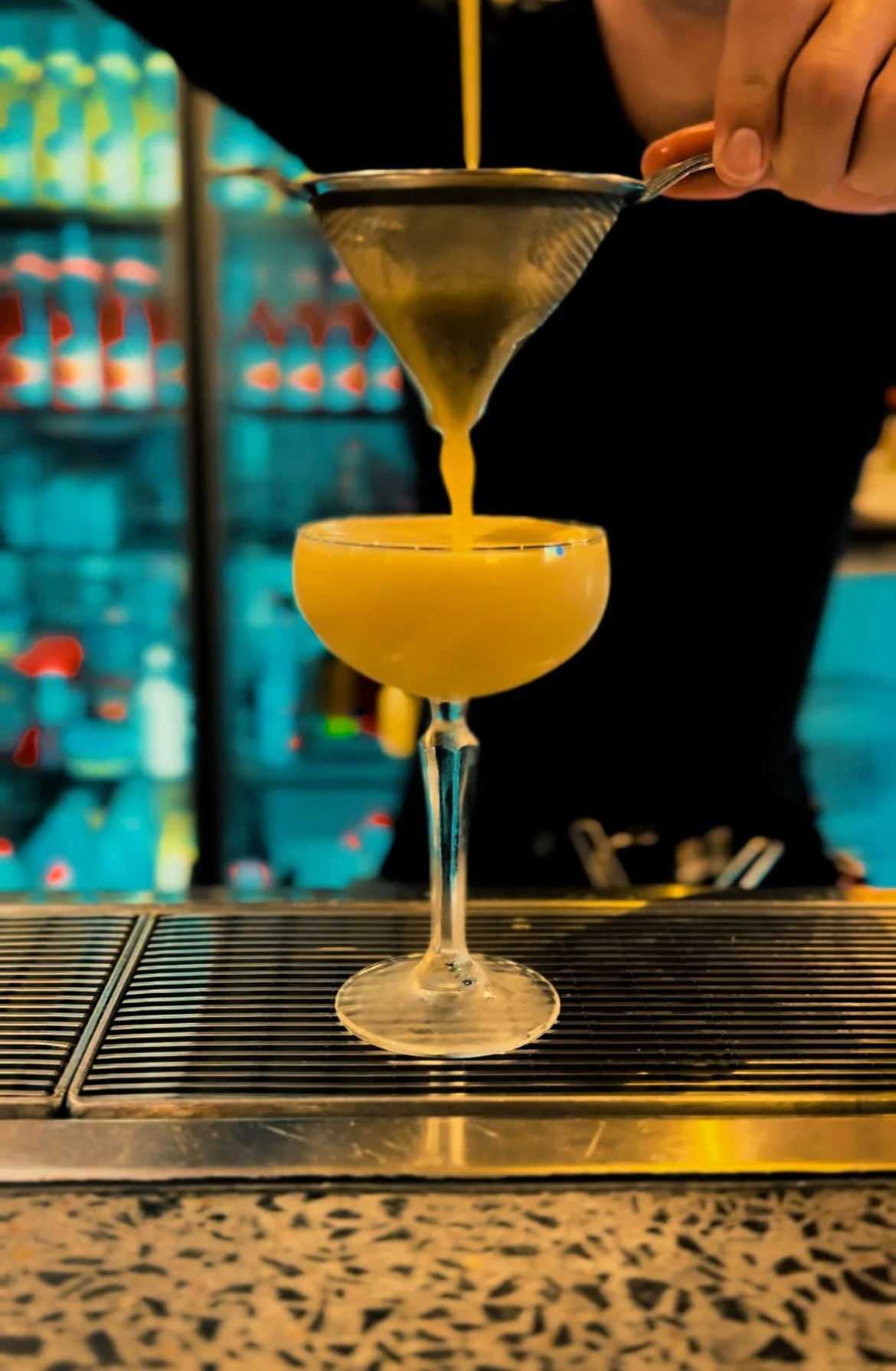 A bartender is pouring a yellow cocktail through a strainer into a coupe glass, creating a layered drink with a yellow top and a creamy white bottom, on a bar counter.