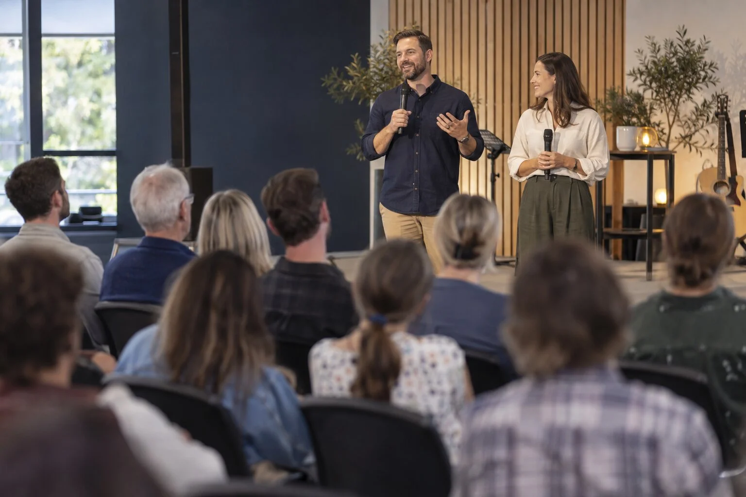 A man and a woman stand on a stage speaking to an audience in a modern, well-lit room with large windows, plants, and musical instruments in the background.
