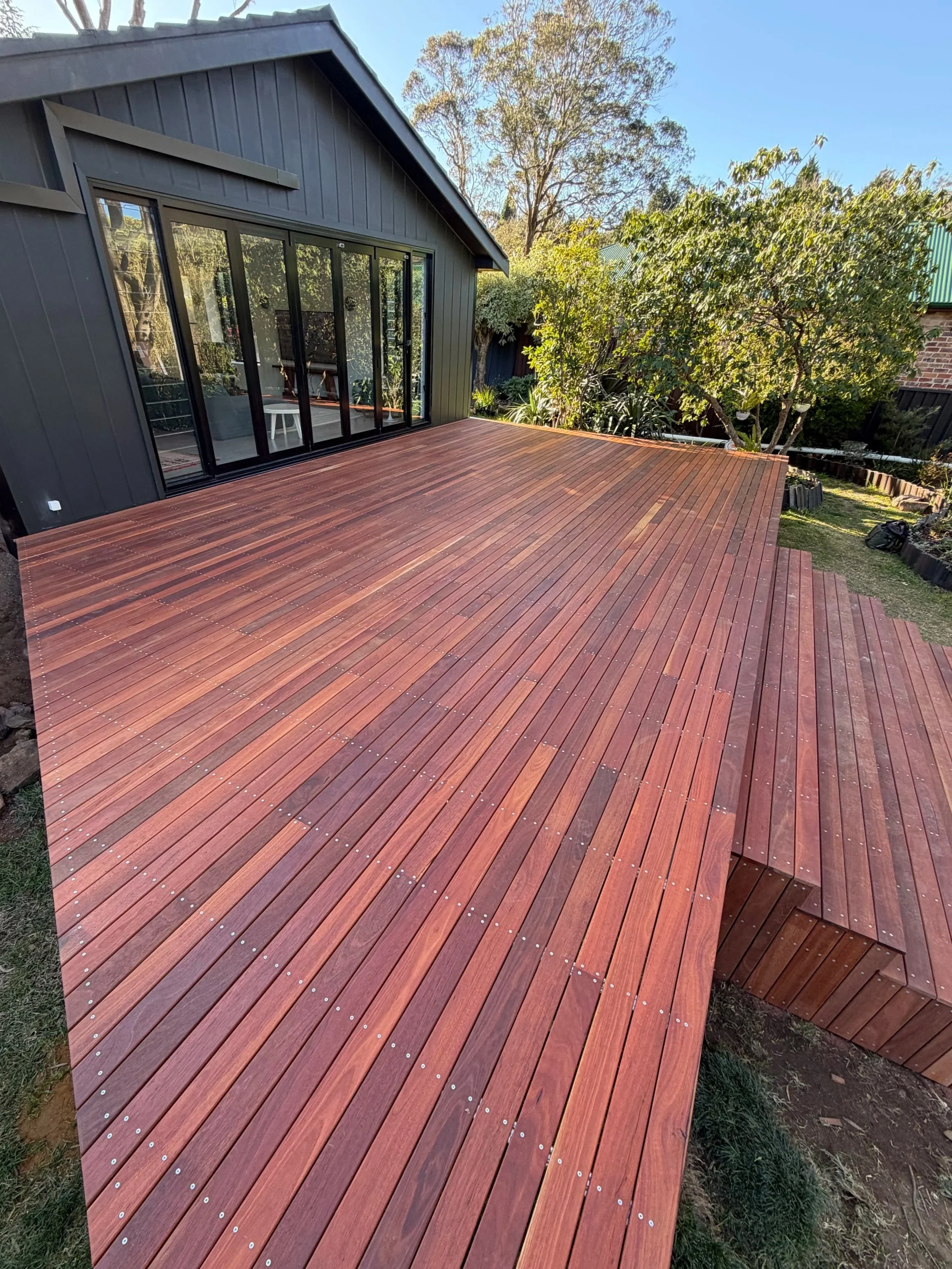 A large wooden deck attached to a dark-colored house with sliding glass doors, surrounded by trees and greenery, in a backyard setting.