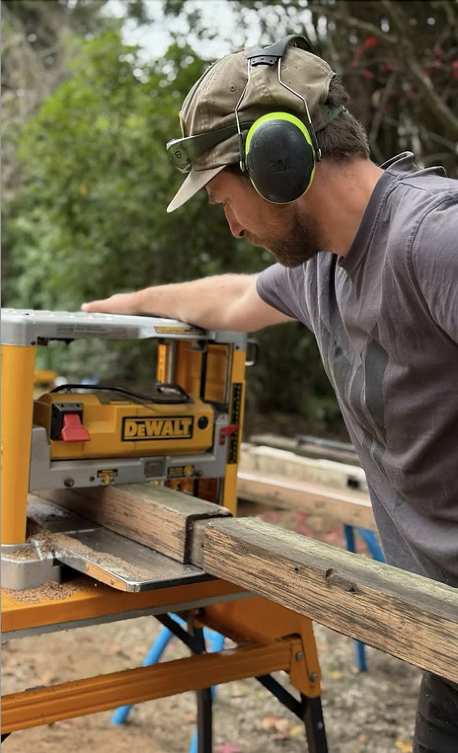 A man wearing a cap, noise-canceling earmuffs, and a gray shirt working with a yellow DeWalt power planer on a piece of wood outdoors.