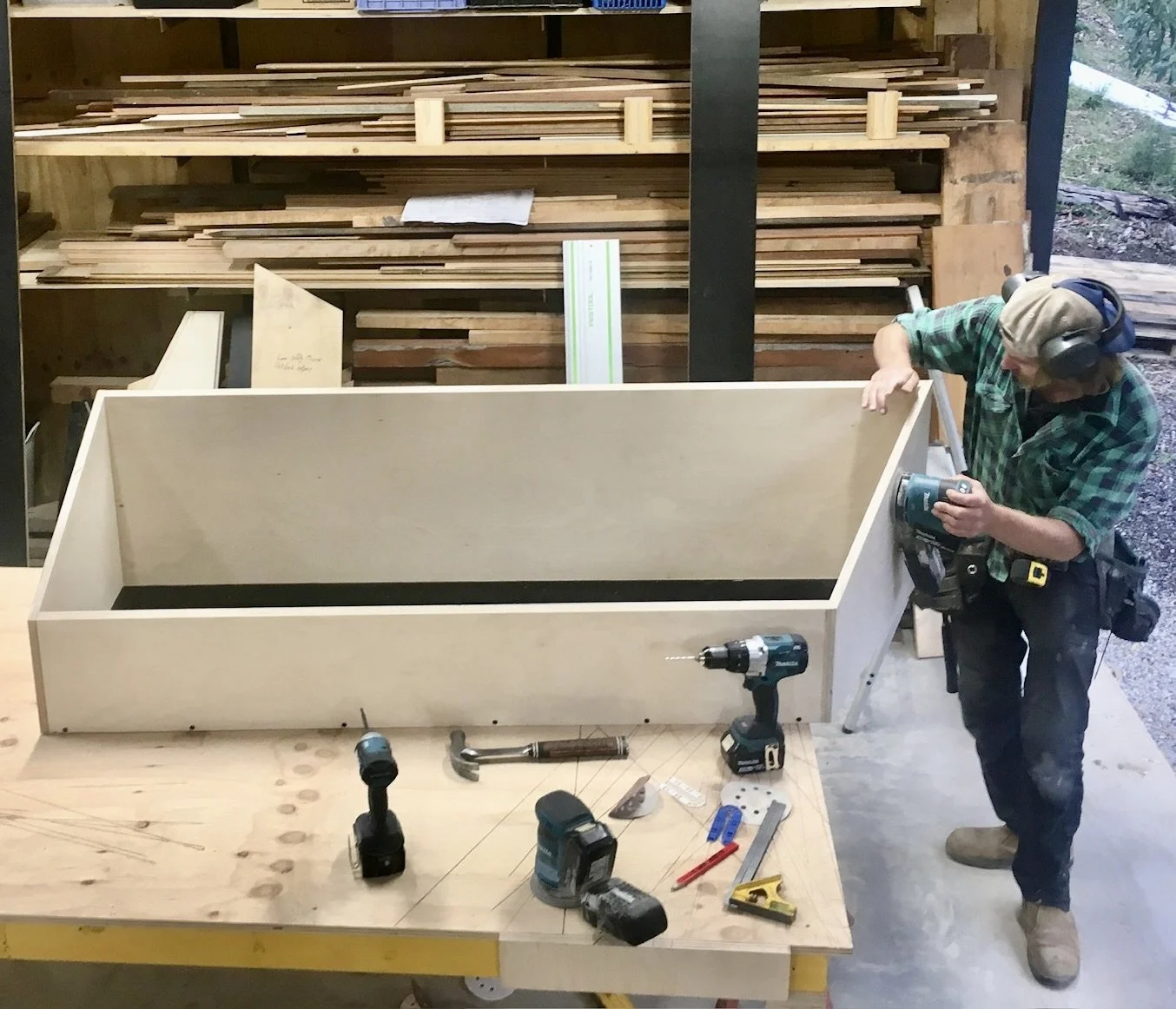 A person working on building a large wooden box in a woodworking shop. There are power tools and hand tools on the table, and shelves filled with various pieces of wood in the background.