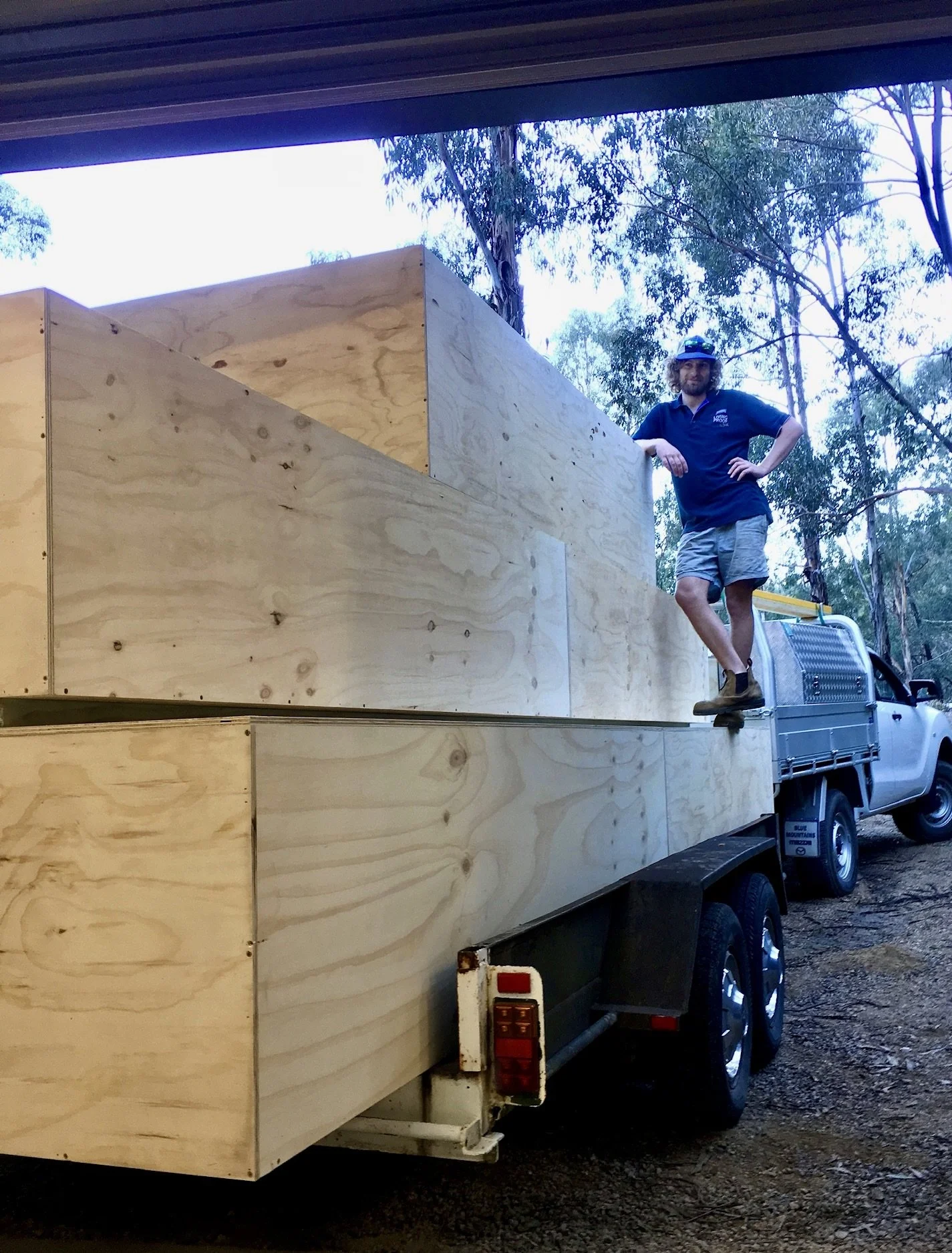 A man standing on the side of a flatbed truck, which is loaded with unfinished wooden boxes, in an outdoor wooded area.