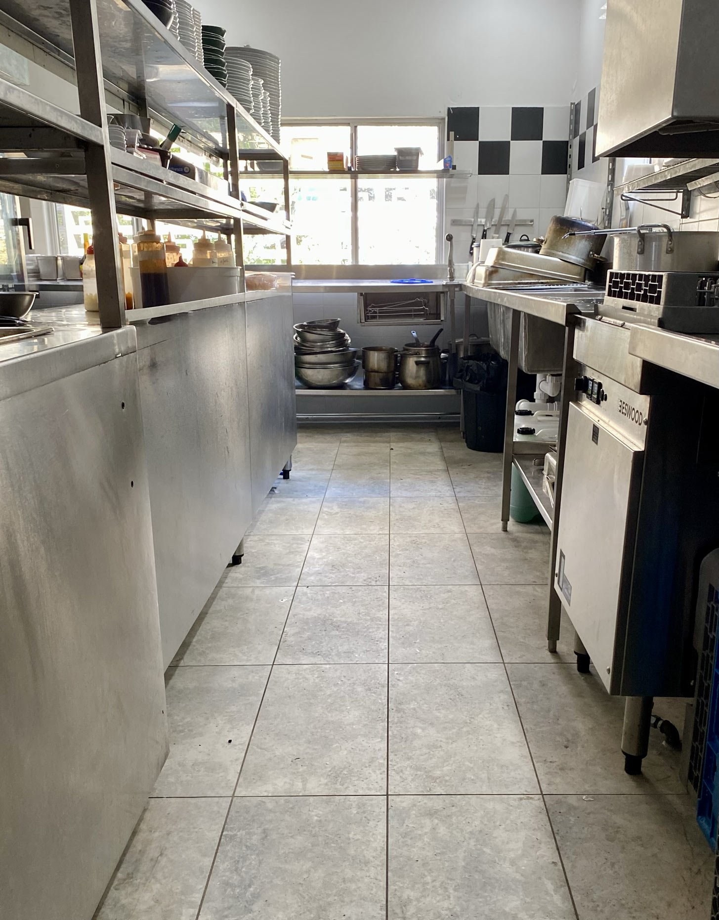 A commercial kitchen with metal shelving on the left holding plates, bowls, and sauce bottles. In the back, there is a window with sunlight coming through, and below it a sink with a faucet. On the right, a stainless steel counter with chafing dishes and kitchen knives on the wall. The floor is tiled in light gray.