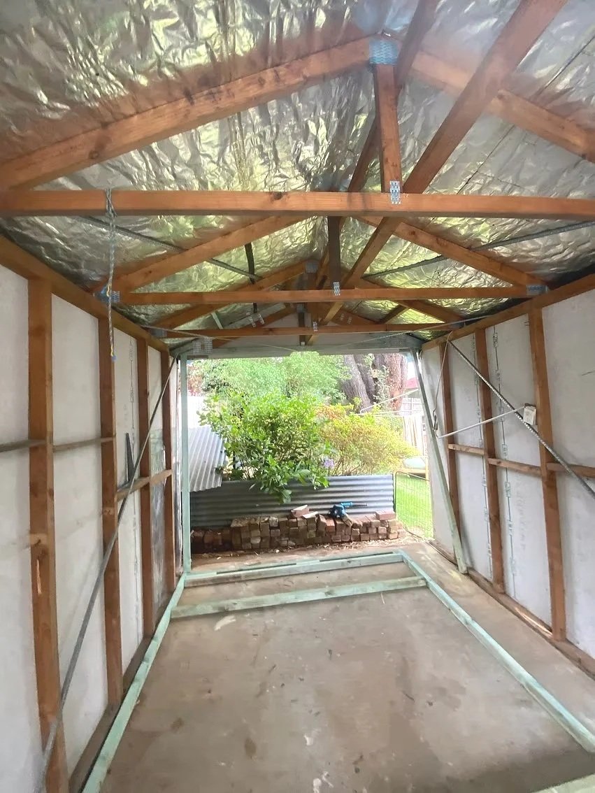 View of a partially built outdoor structure with wooden framing, reflective insulation on the roof, and a concrete floor, with a garden and trees visible in the background.