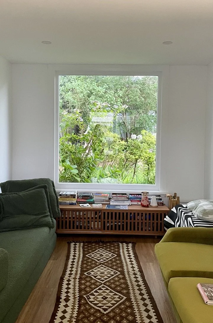Living room with big window showing green garden outside, green sofa on the left, yellow chair on the right, wooden pet crate with books on top, patterned rug on the floor, and various items on the window sill.
