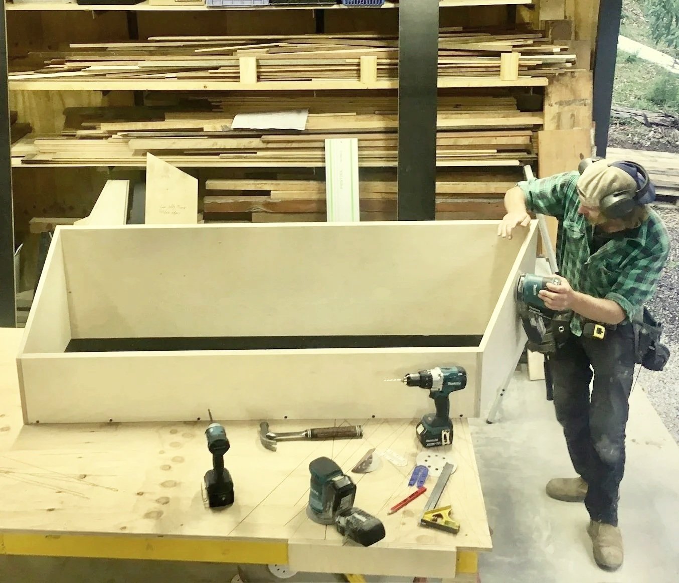 Man with safety headphones working on assembling a wooden structure in a woodworking shop, with tools and wood materials around him.