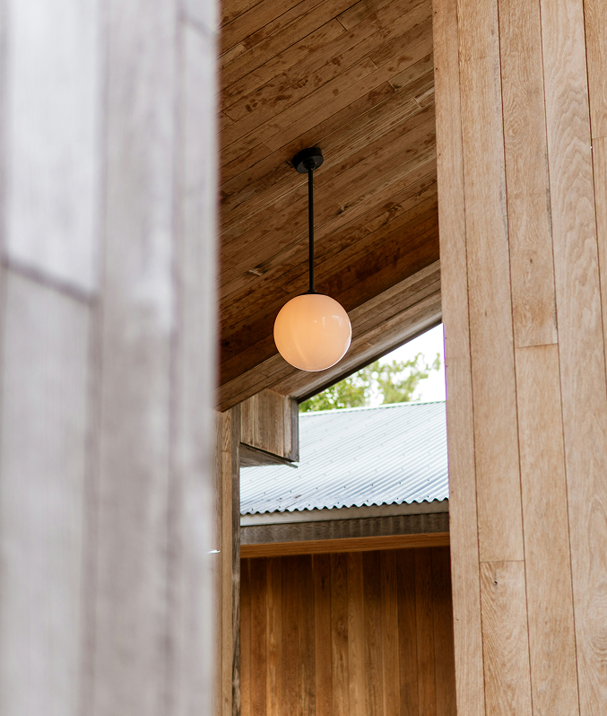 A round, white pendant light hanging from a black cord against a wooden ceiling and walls, with a partial view of a metal roof outside a window.