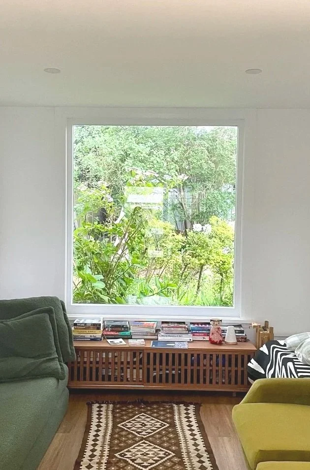 Living room with a large window overlooking green foliage, bookshelf with books, a green sofa, and a yellow chair.