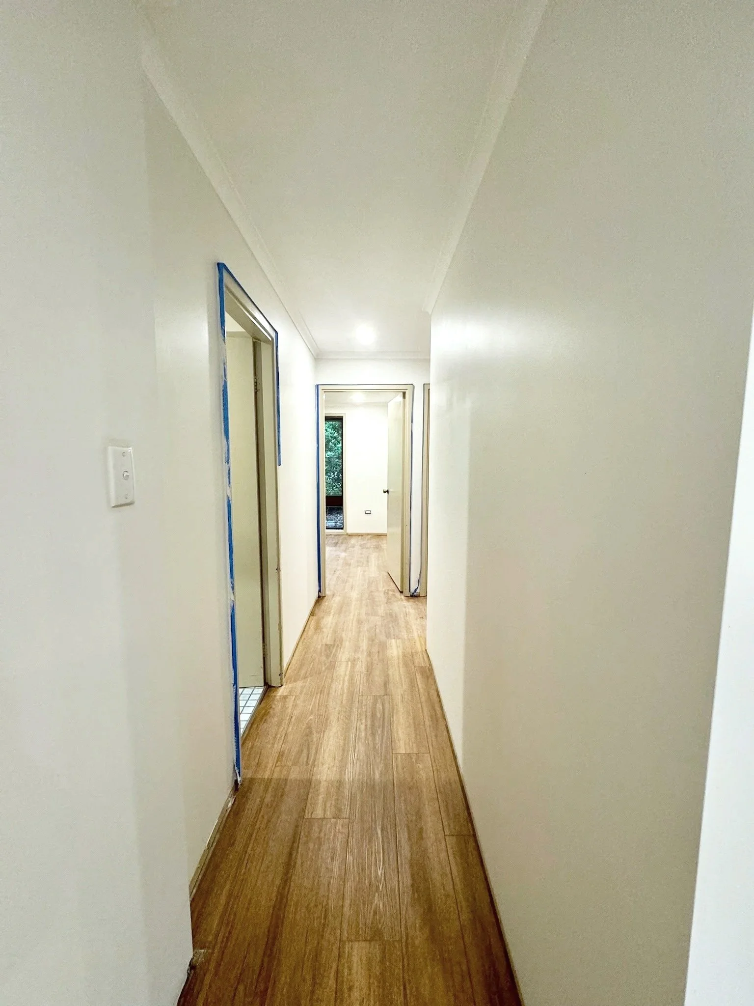 A narrow hallway with white walls, wooden flooring, and multiple doorways leading to rooms. One door on the left has painter's tape around its frame. Natural light is visible at the end of the hall.