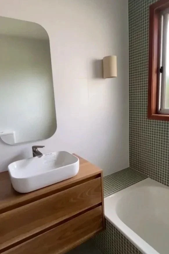 Bathroom with a wooden vanity, a white vessel sink, a mirror, a wall-mounted light fixture, small green tiles around the bathtub and window, and a window with wooden trim.