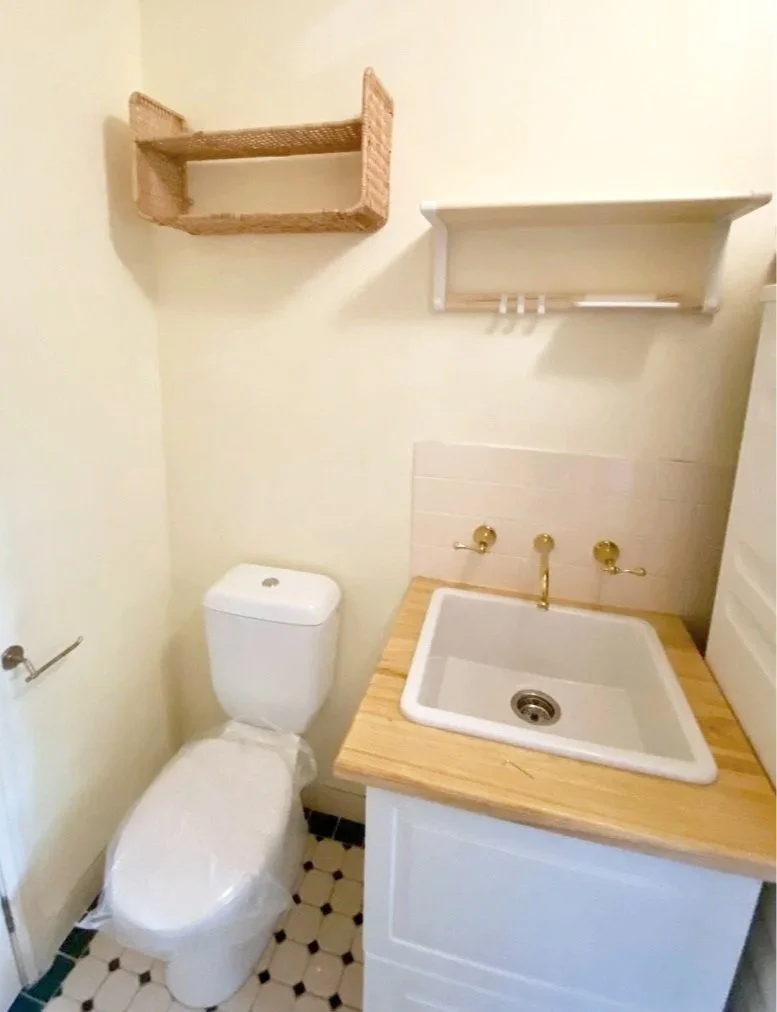 Small bathroom with a toilet, a wooden sink with brass fixtures, and wall-mounted shelves, one wicker and one painted white.