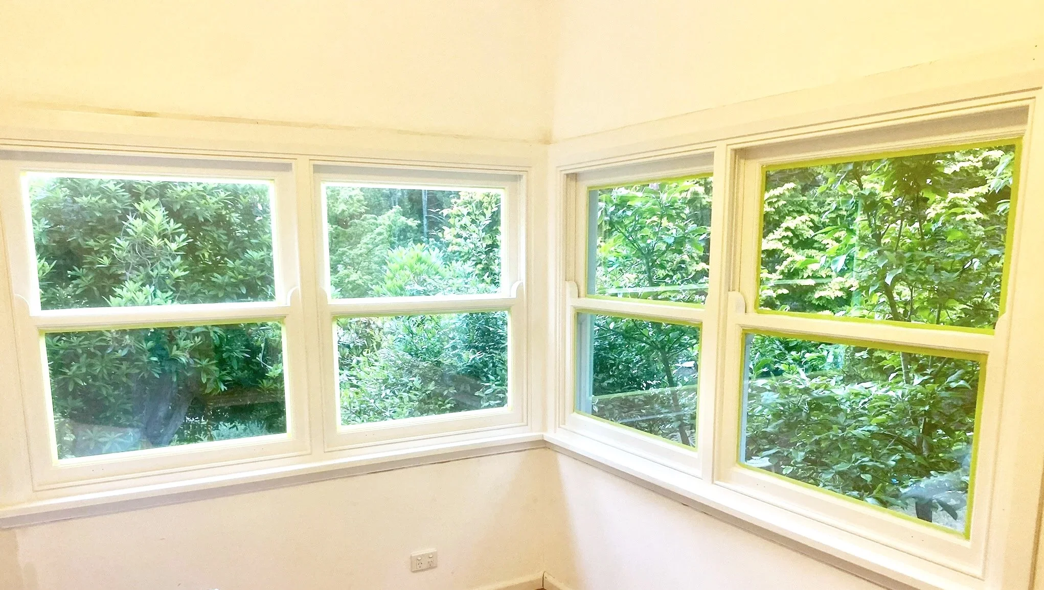 A corner room with three large windows showing lush green trees outside.