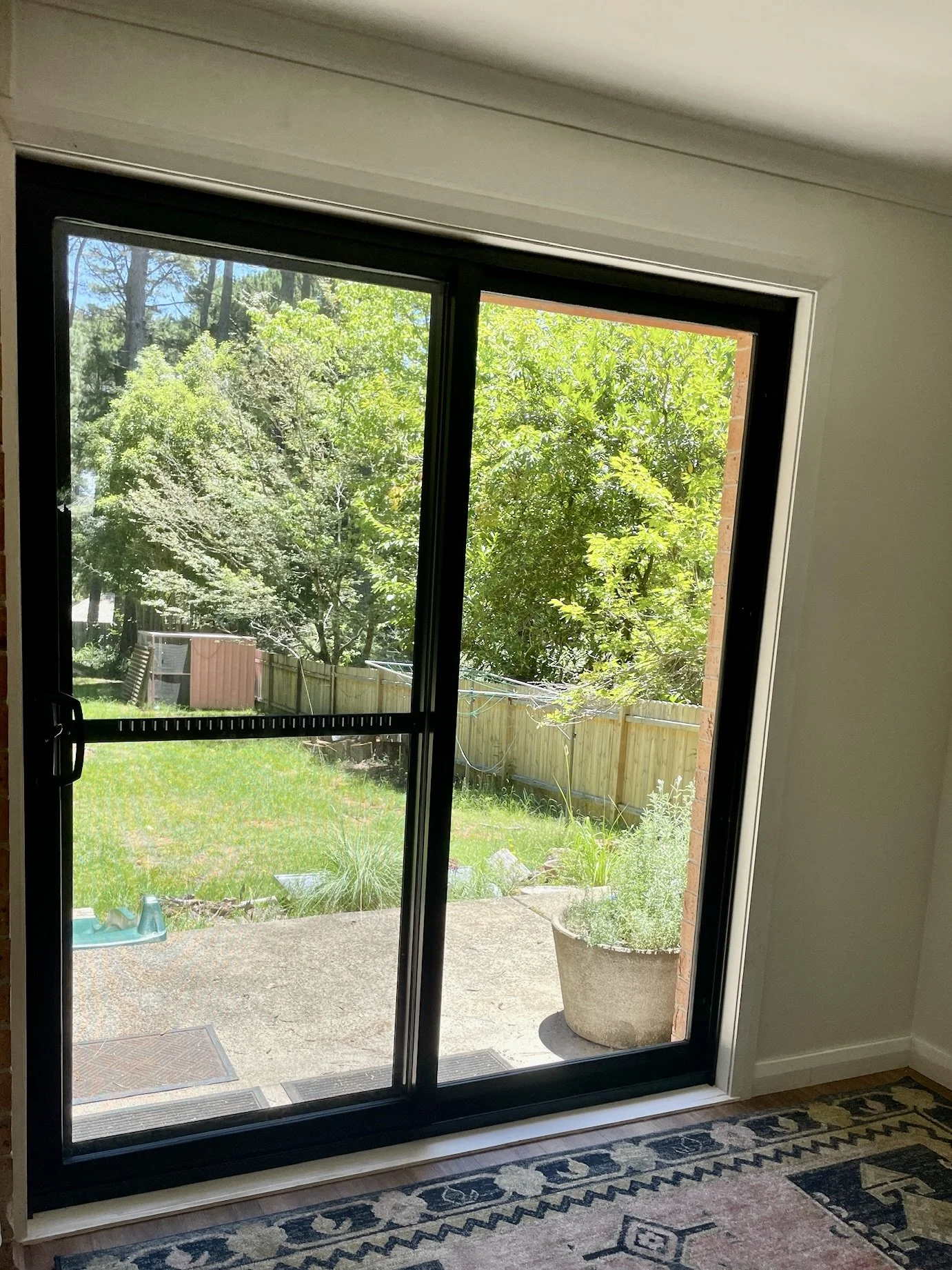 A view through a sliding glass door opening to a backyard with green grass, trees, and a wooden fence. Part of the interior, including a patterned rug on the floor, is visible.