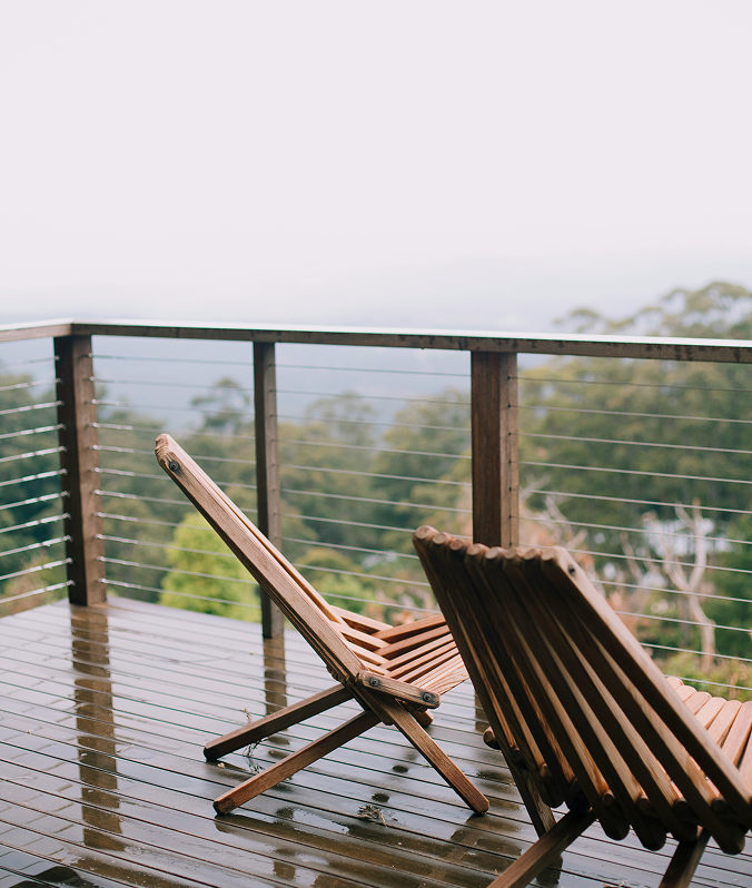 Two wooden lounge chairs on a wet deck with a view of foggy forested hills in the background.
