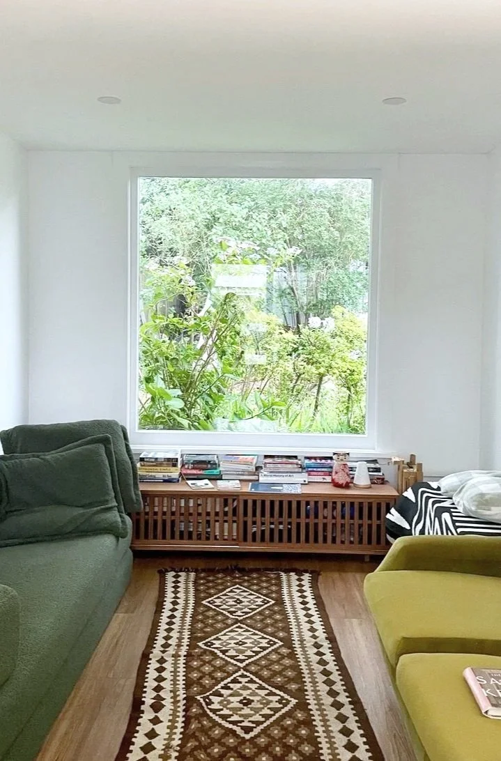 A cozy living room with a large window overlooking a lush, green garden. There are two green armchairs, a wooden radiator cover filled with books, a patterned rug on a wooden floor, and some pillows and decorative items on the furniture.
